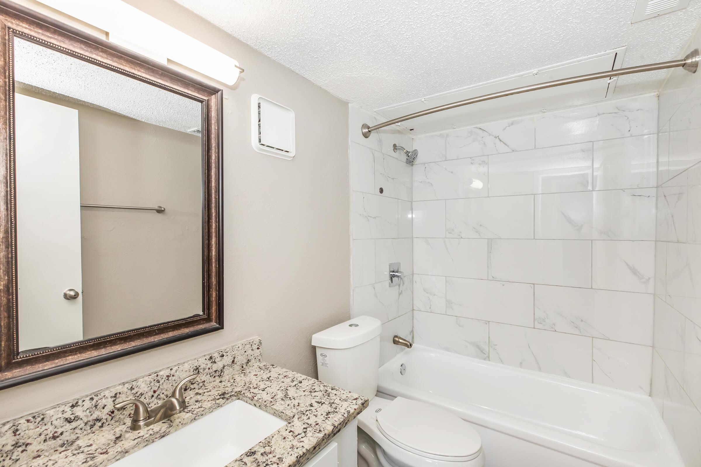 A modern bathroom featuring a white bathtub with a shower, a toilet, and a sink with a granite countertop. There is a large mirror above the sink and stylish light fixtures. The walls are adorned with white marble tiles, giving the space a clean and elegant appearance.