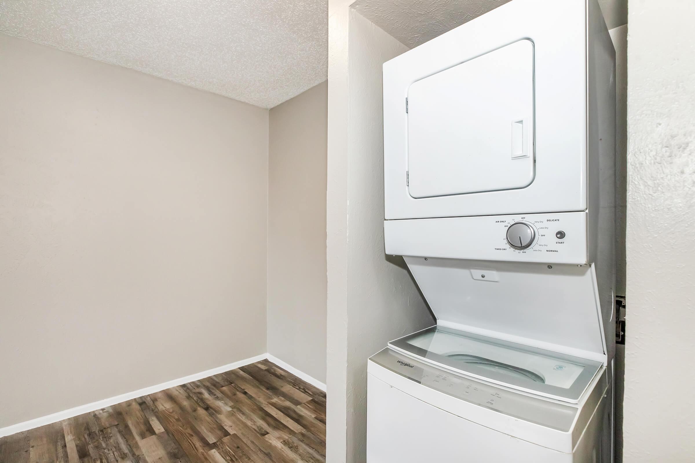 A compact laundry area featuring a stacked washer and dryer against a light beige wall. The floor is covered with wooden planks, and there is an empty space or room visible on the left. The overall ambiance is clean and functional, suitable for laundry tasks.