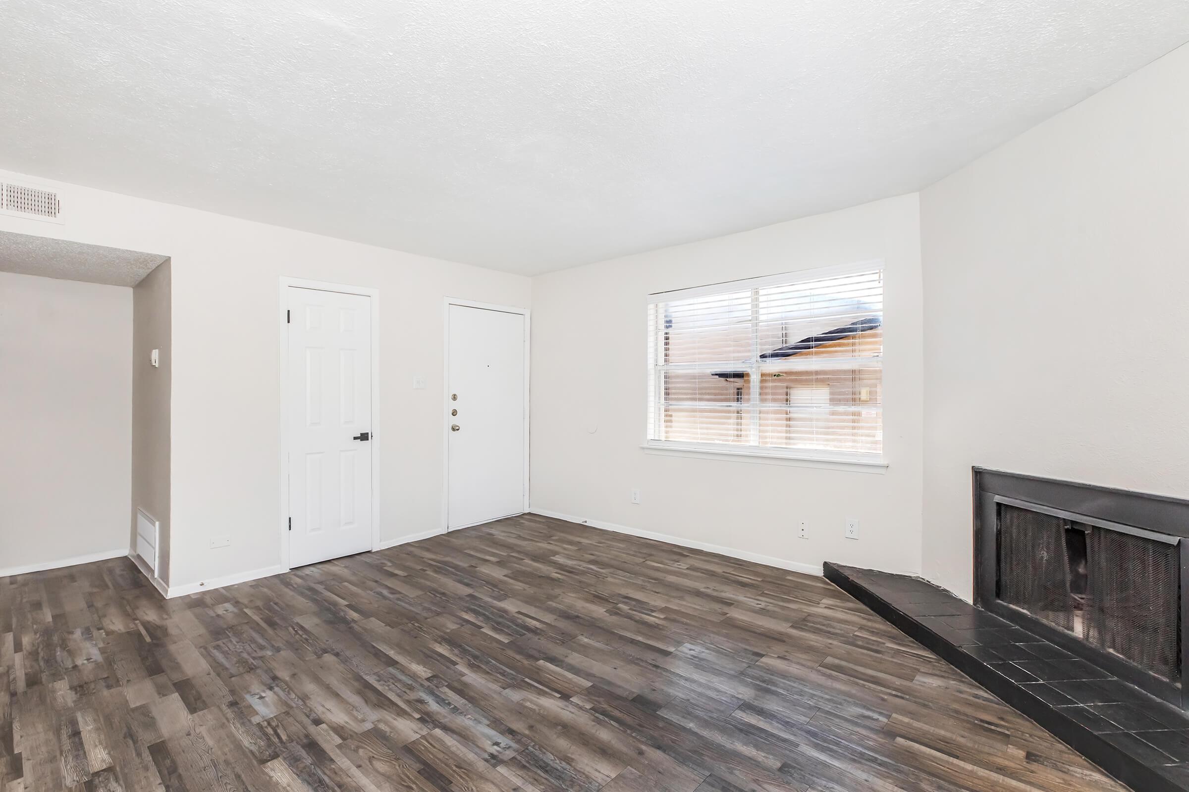A well-lit living room featuring light-colored walls, a large window with blinds, light wood flooring, and a fireplace. The room has a simple door and a spacious feel, with an uncluttered design.