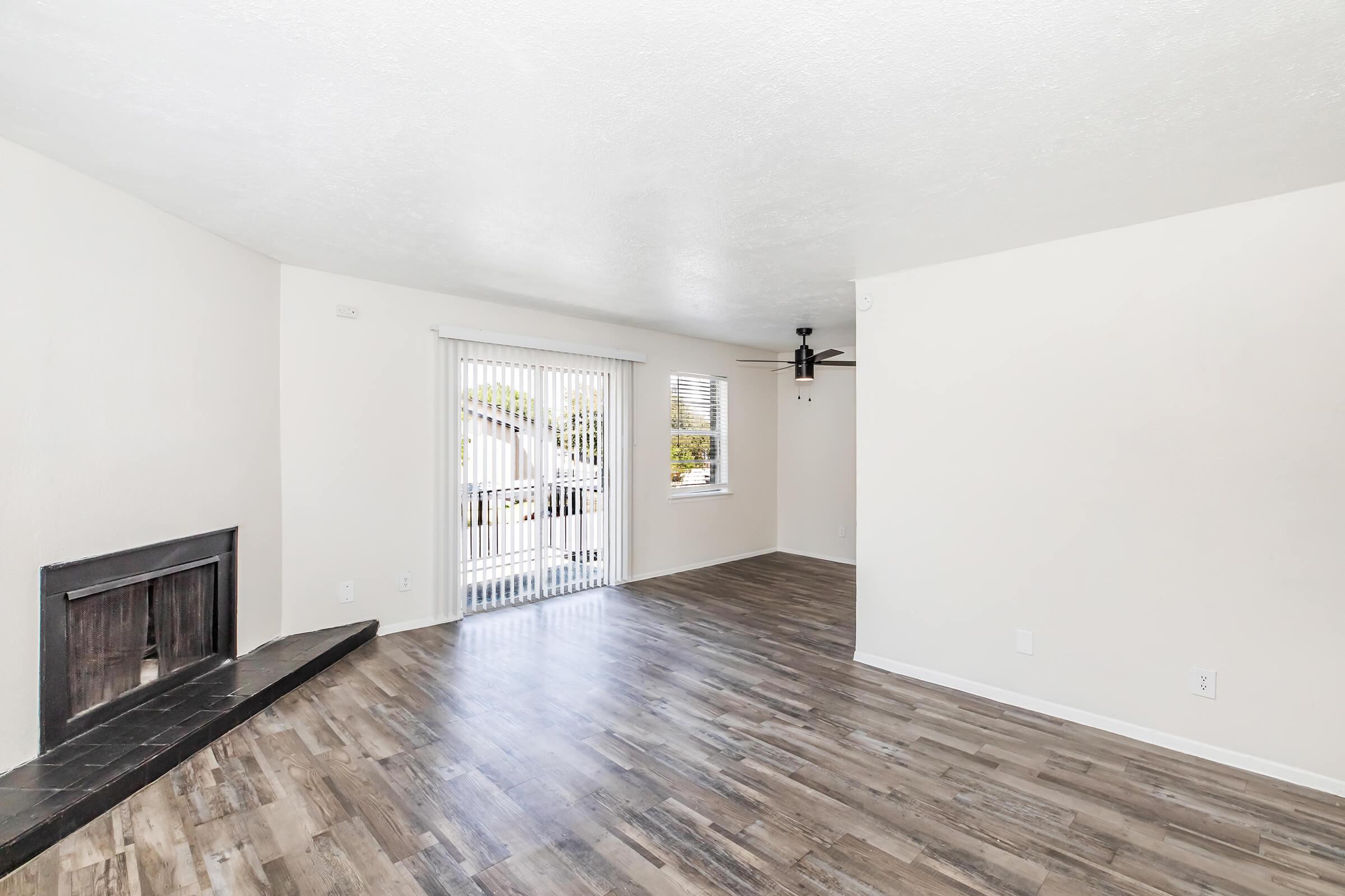  spacious living room with light-colored walls and wood-like flooring. It features a large window with vertical blinds, a ceiling fan, and a fireplace with a black hearth. The room has an open layout, allowing natural light to fill the space.