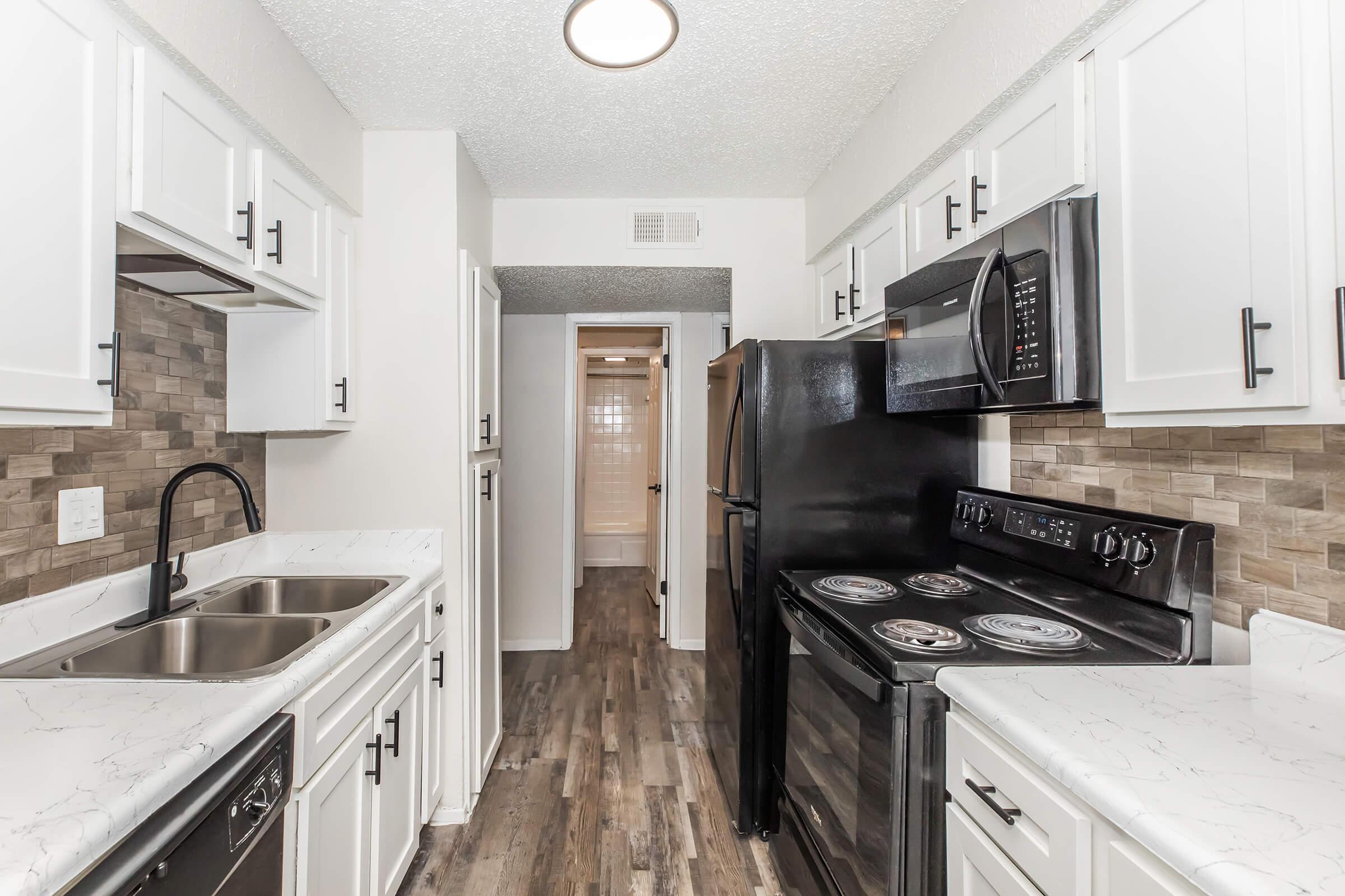Modern kitchen featuring white cabinetry, dual sinks, a black stove, and refrigerator. Gray and beige tiled backsplash adds texture. Light-colored wood flooring complements the space, which opens to a hallway leading to other rooms. Bright overhead lighting enhances the clean, contemporary design.