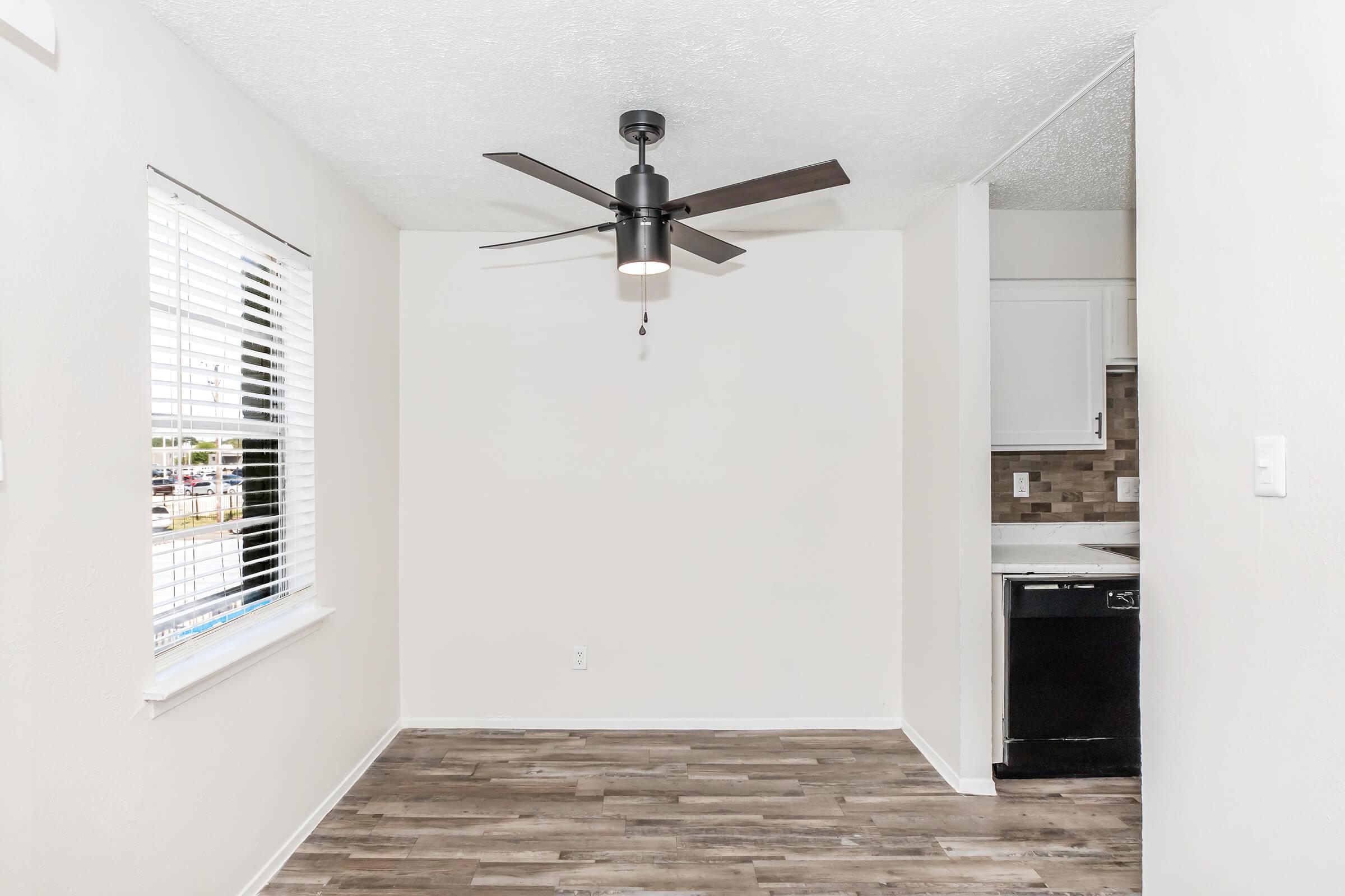 A bright, airy room featuring a ceiling fan and large window with blinds. The walls are painted white, and the flooring is light-colored wood. In the background, there's a glimpse of a kitchen area with dark appliances and a modern backsplash. The overall atmosphere is clean and inviting.