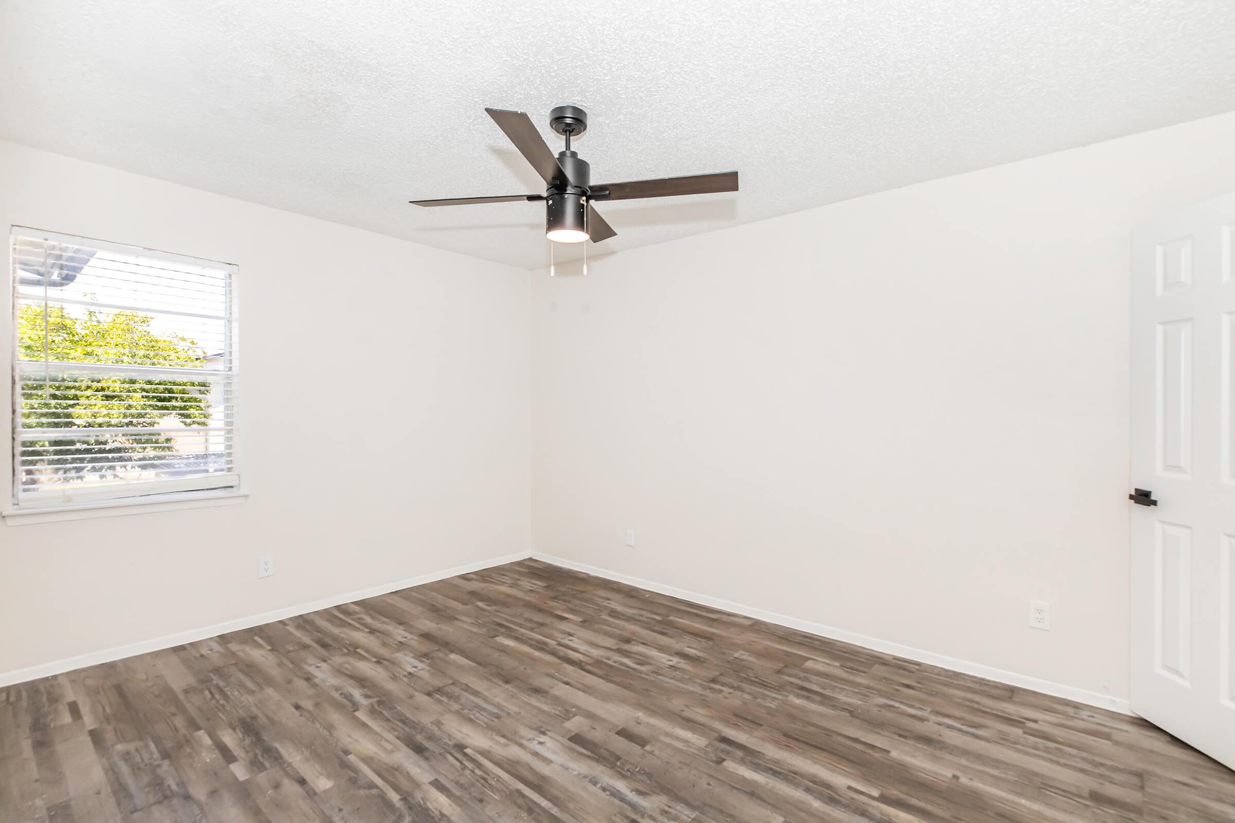 A bright, empty room with light-colored walls and a ceiling fan. The flooring is wood-like, and there's a window with blinds allowing natural light in. The overall space is clean and minimalistic, suitable for various uses. A closed white door is visible on the right.