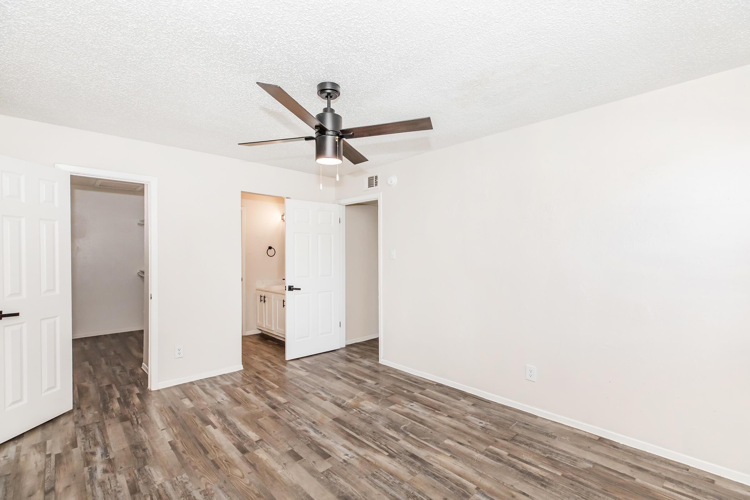 A bright and spacious interior room with light beige walls and a textured ceiling. The floor features wood-like laminate planks. A ceiling fan with a modern design hangs from the ceiling. Two white doors lead to adjacent rooms, and a small area with a sink is visible in the background.
