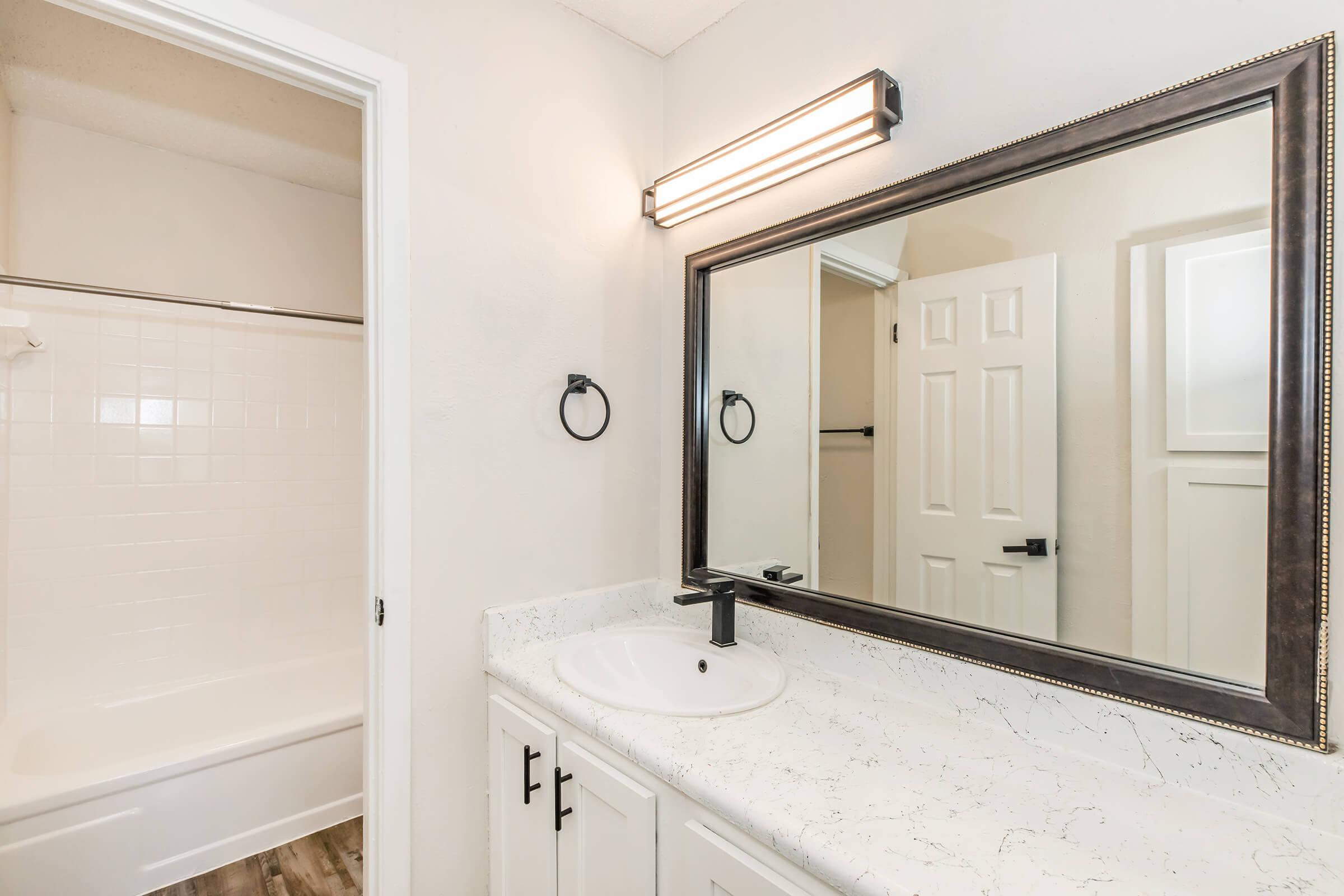 Modern bathroom featuring a stylish sink with a dark faucet, a large mirror with a black frame, and wall-mounted lighting. The space includes a white bathtub and a shower area, with light-colored walls and cabinetry, creating a clean and contemporary look.
