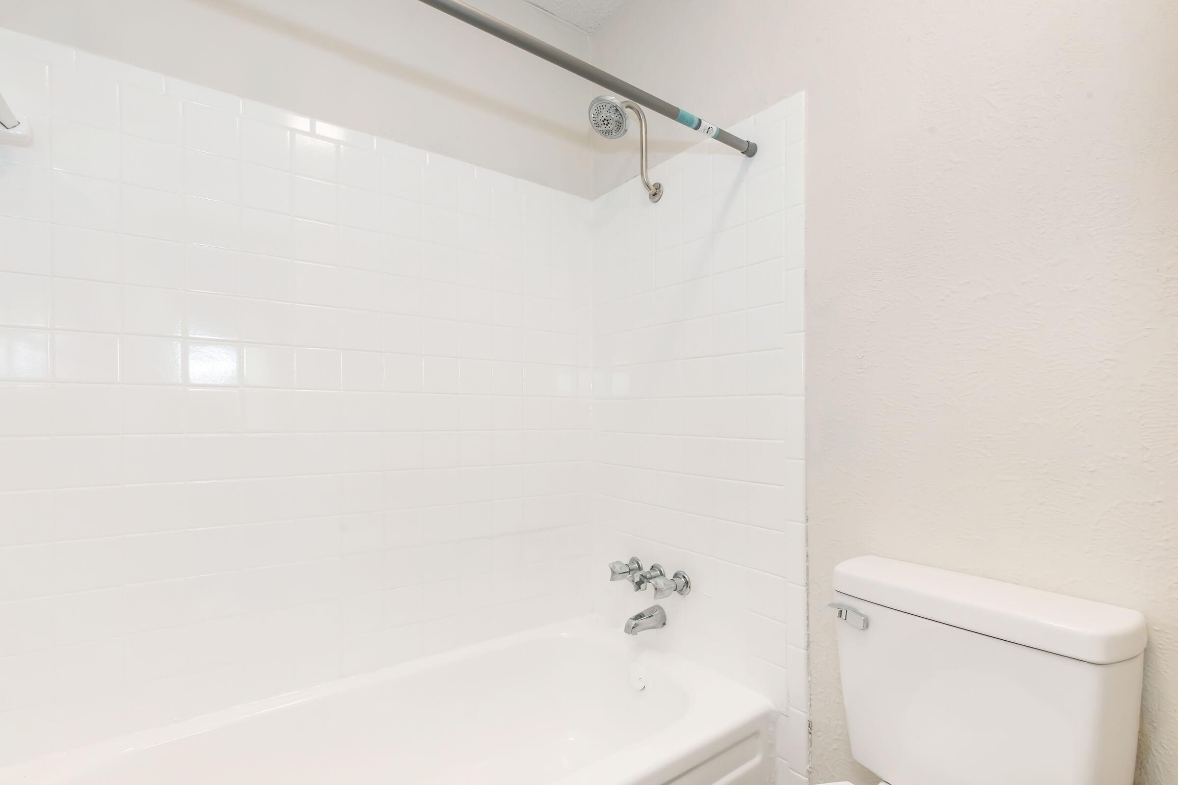A clean, bright bathroom featuring a white tiled shower area with a bathtub, a chrome showerhead, and a toilet next to it. The walls are painted a light color, enhancing the spacious feel of the room.