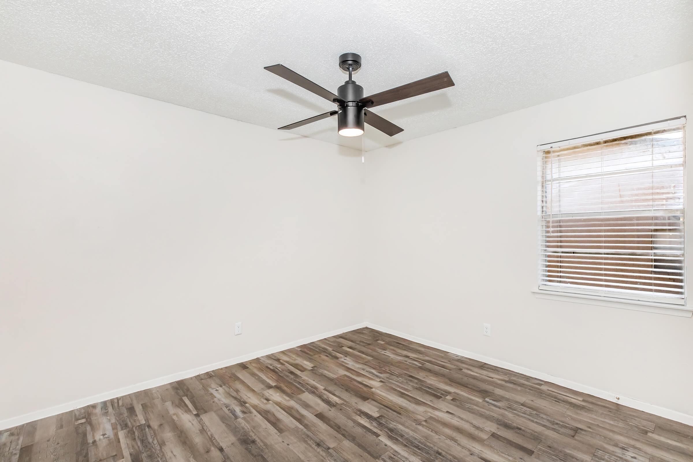 A bright, empty room featuring light-colored walls and a ceiling fan with dark blades. The floor has light brown wooden planks. A window with blinds allows natural light to enter, creating a spacious and minimalistic atmosphere.