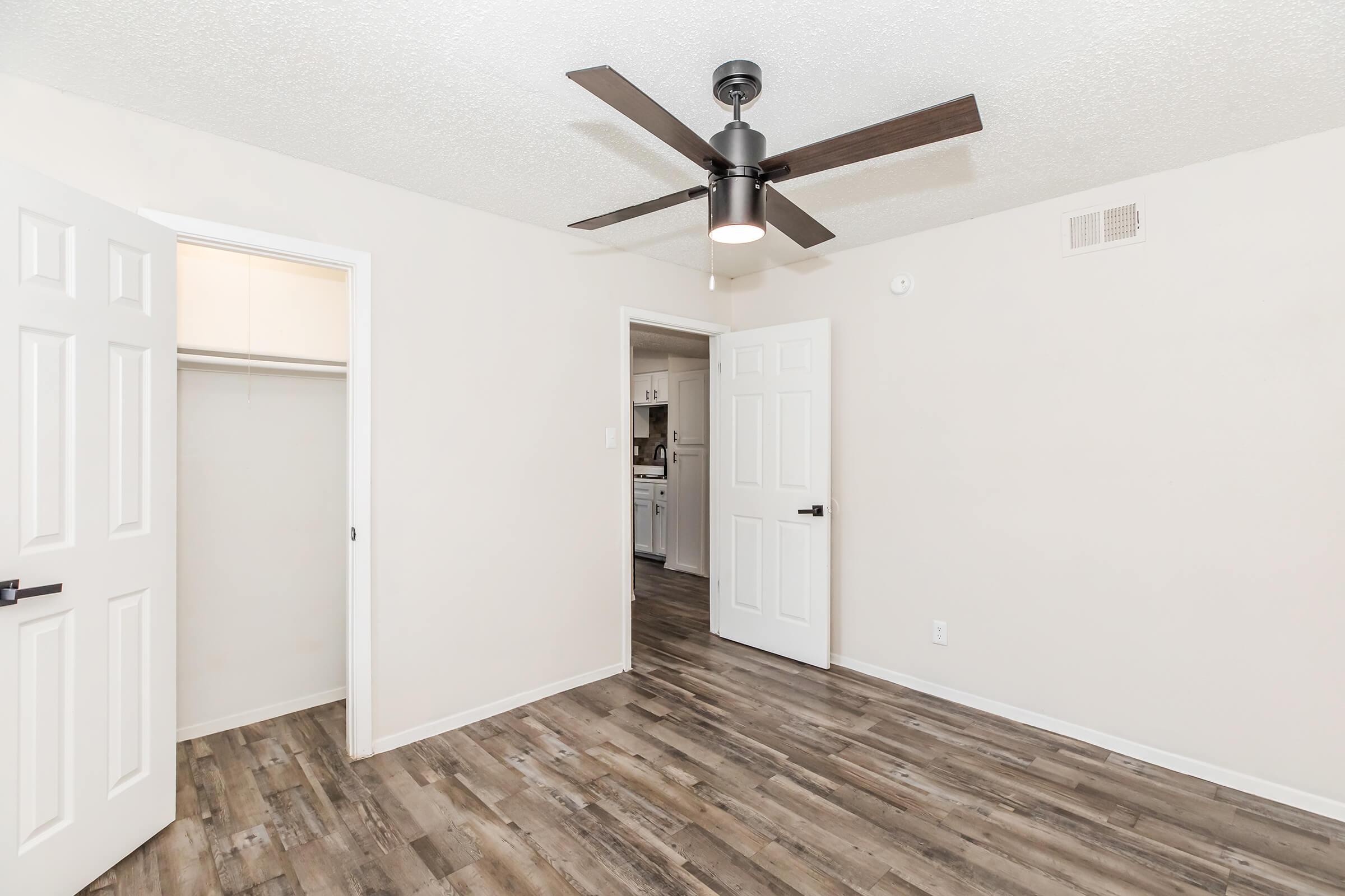 A spacious room featuring light-colored walls and wood-like flooring. It includes a ceiling fan and two open doors leading to a closet and an adjacent kitchen area. Natural light fills the space, creating a bright and inviting atmosphere.