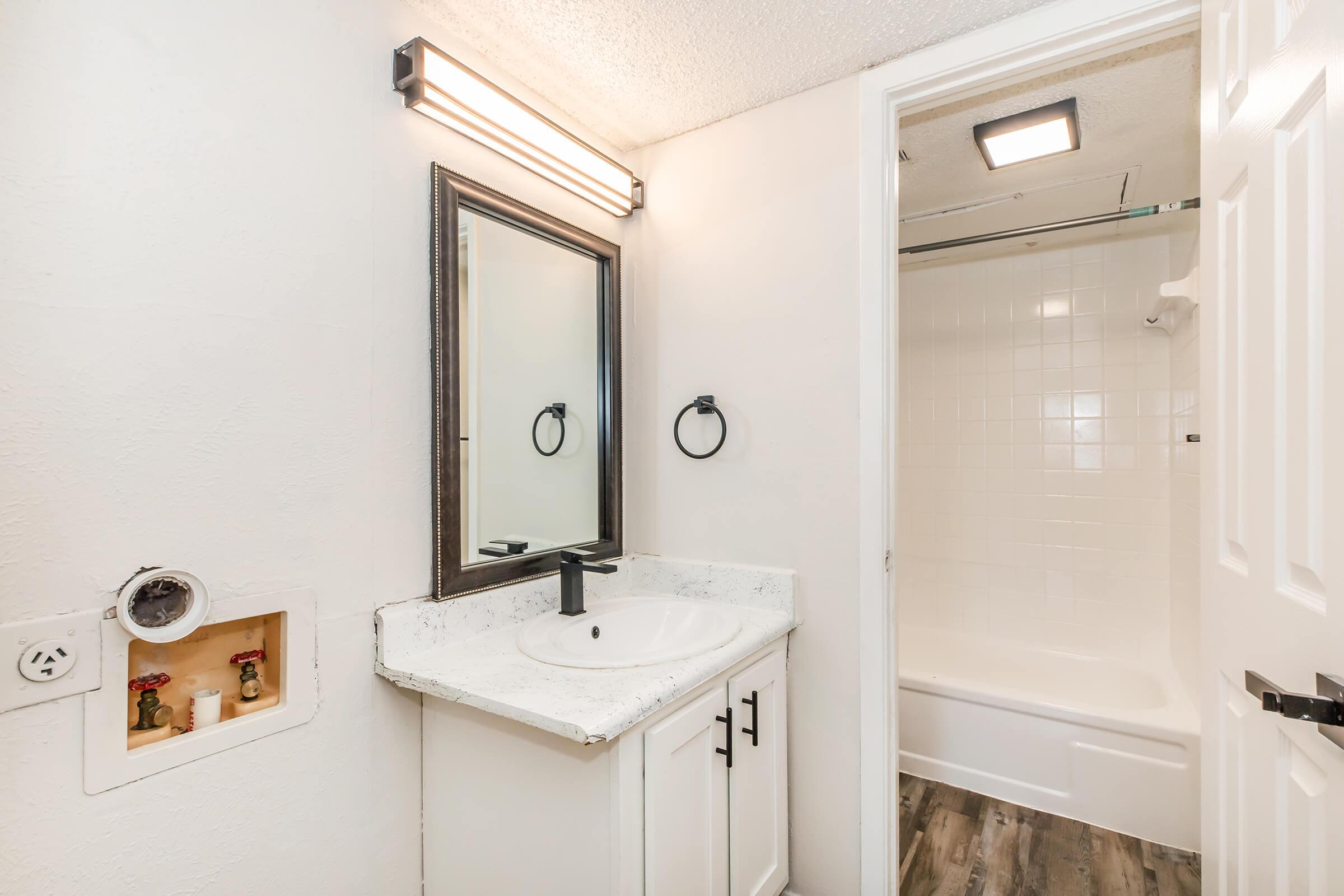 A bright and modern bathroom featuring a white vanity with a sink and a large mirror above. There is a towel ring next to the mirror, and behind the sink is a small wall opening. To the right, a door leads to a clean, tiled shower and bathtub area. The flooring is wood-like laminate, contributing to the contemporary look.