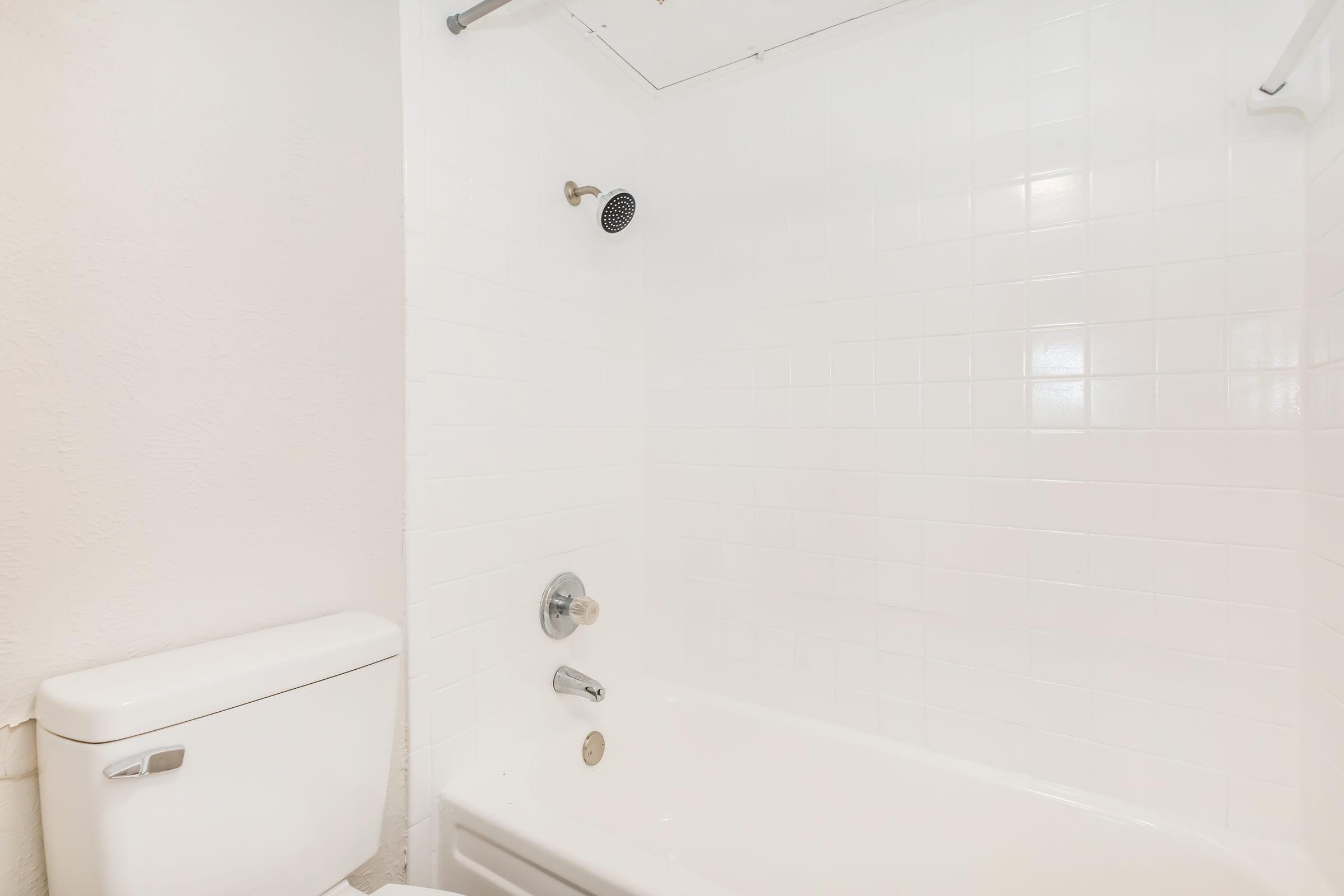 A clean and modern bathroom featuring a white bathtub and shower with a wall-mounted showerhead, alongside a matching white toilet. The walls are textured and light-colored, enhancing the bright and airy feel of the space.