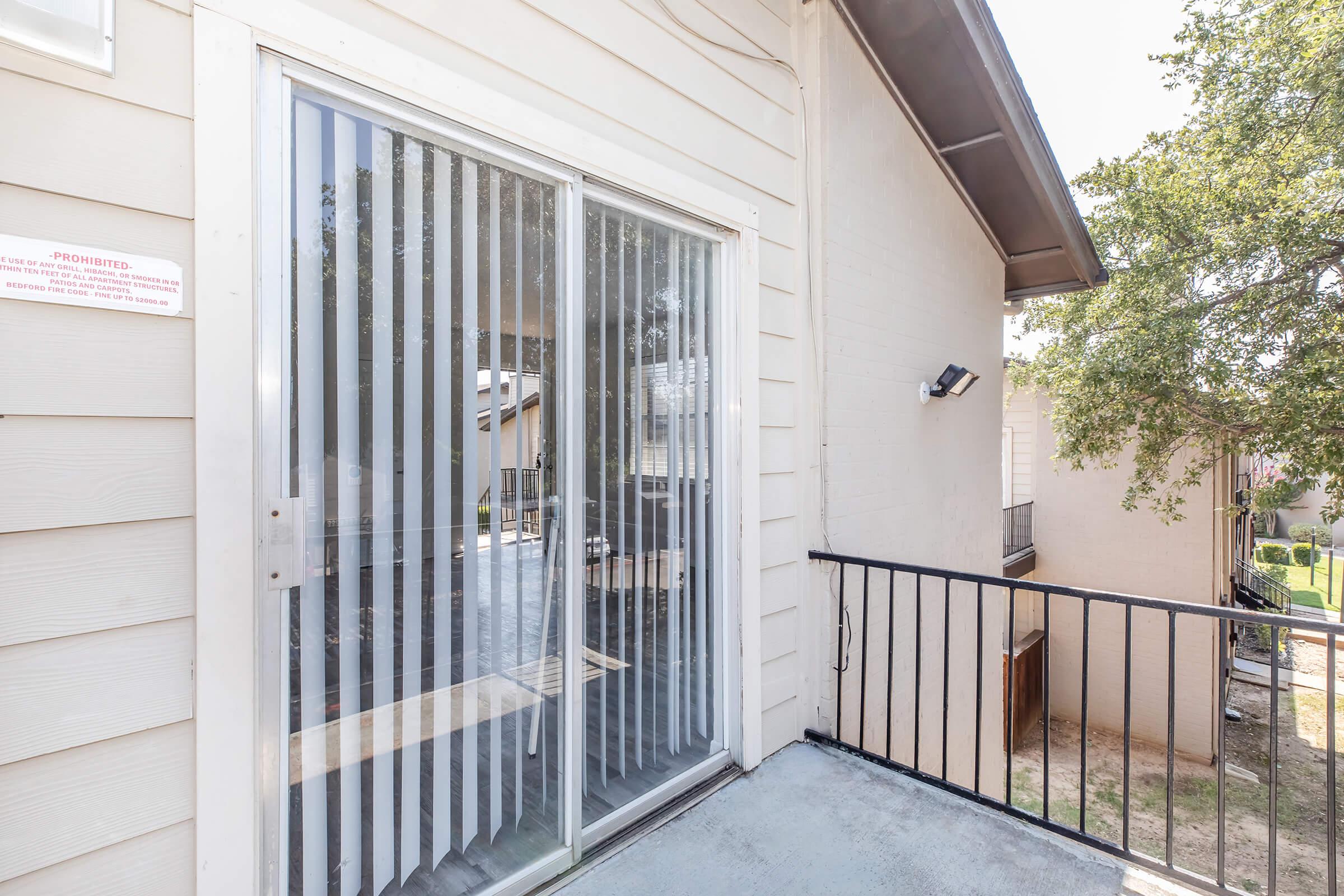 A sliding glass door with vertical blinds leads to a balcony area. The exterior features light-colored siding and a railing. A small tree is visible in the foreground, with a view of neighboring buildings and greenery in the background. Bright sunlight illuminates the scene.