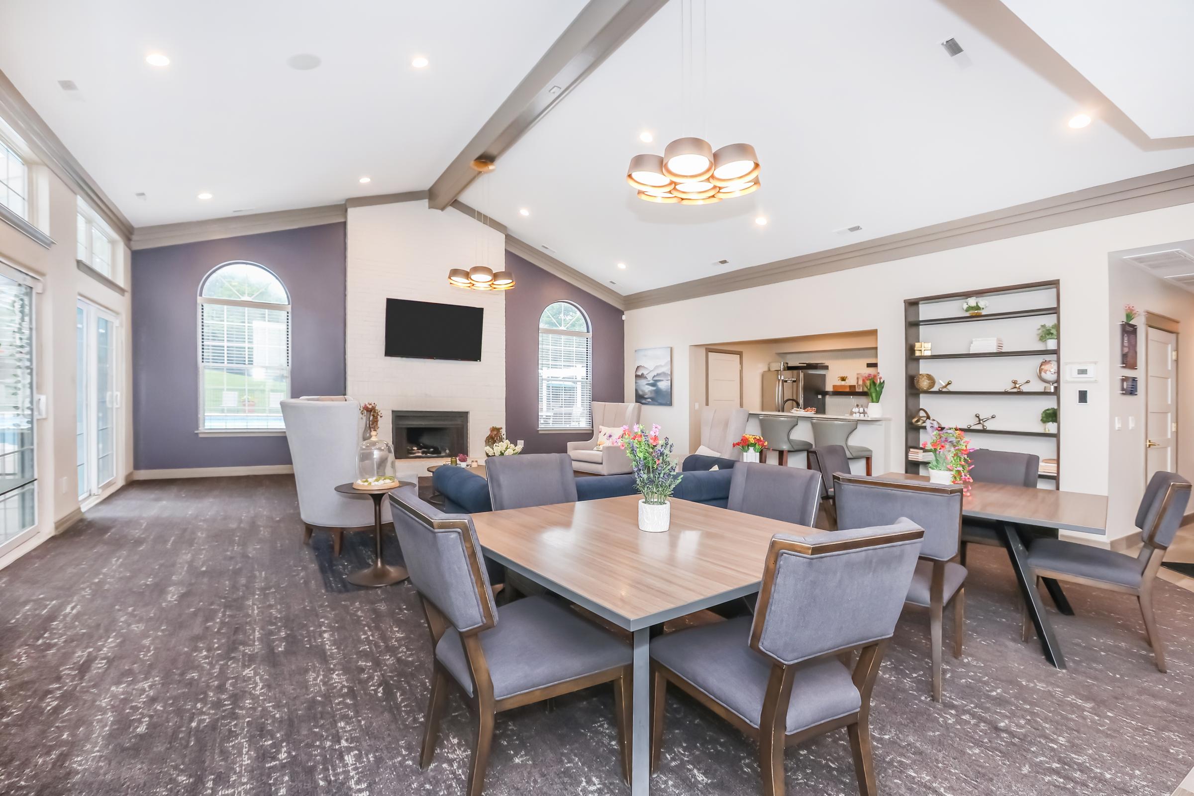A modern dining or community room featuring a large table surrounded by upholstered chairs. The space has large windows, a cozy fireplace, and tasteful decor including flowers on the table. Bright lighting and a stylish ceiling fixture enhance the inviting atmosphere.