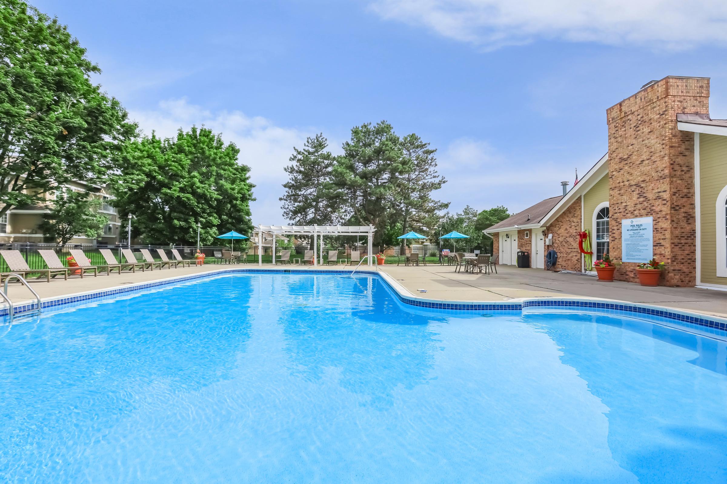 A clear blue swimming pool surrounded by lounge chairs and greenery, with umbrellas providing shade. In the background, there are trees and a building with a brick facade, creating a serene and inviting atmosphere for relaxation and leisure.