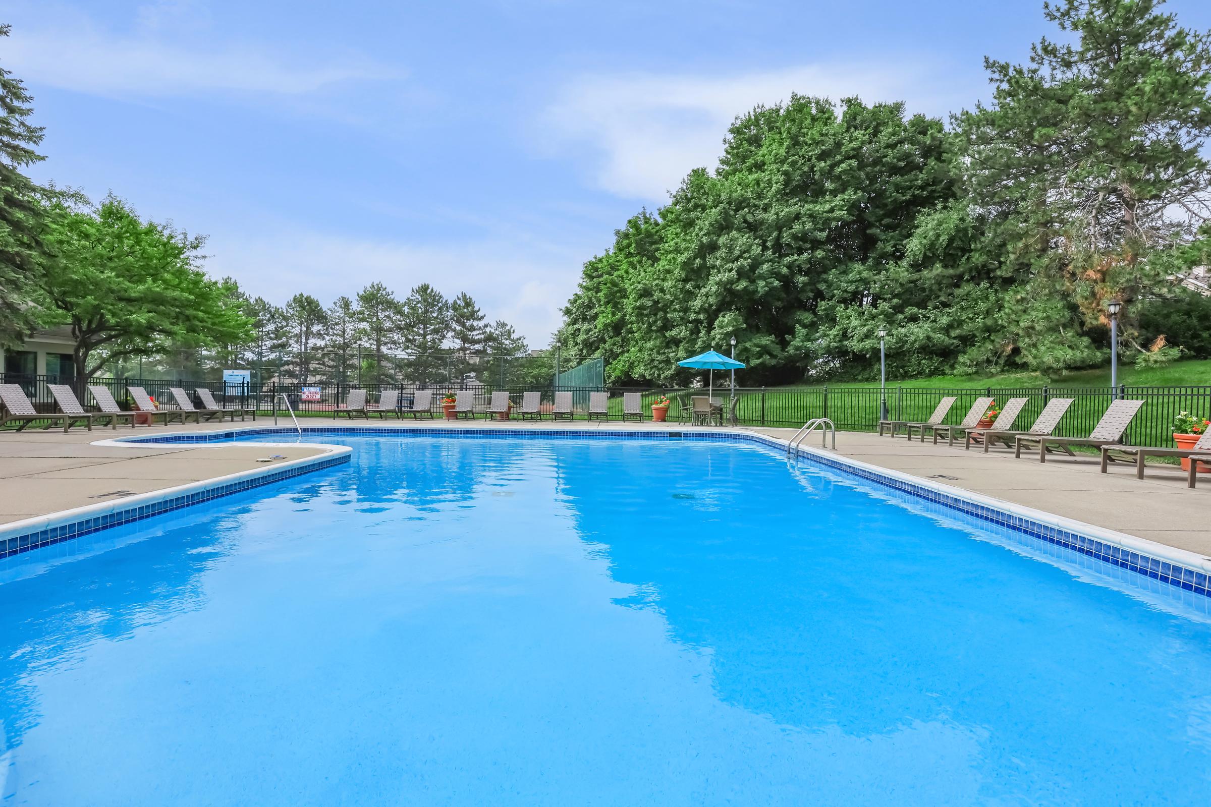 A clear blue swimming pool surrounded by lounge chairs, set against a backdrop of lush green trees and a blue sky. A shaded area with an umbrella adds to the inviting atmosphere, making it a perfect spot for relaxation and leisure.