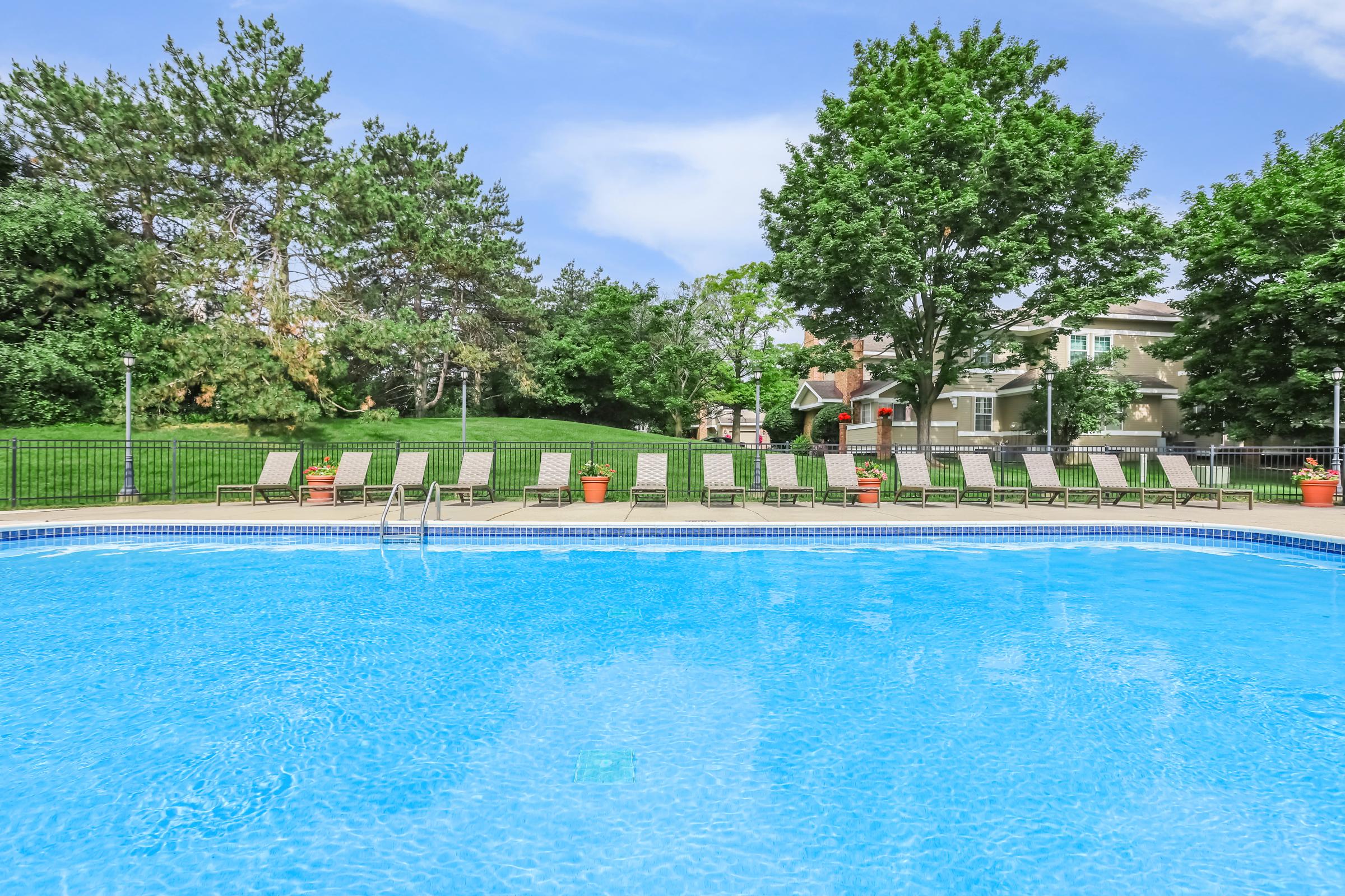 A clear blue swimming pool surrounded by lounge chairs, with green trees and a grassy area in the background. The poolside area is well-maintained, featuring potted plants and a fence, creating a relaxing outdoor setting.