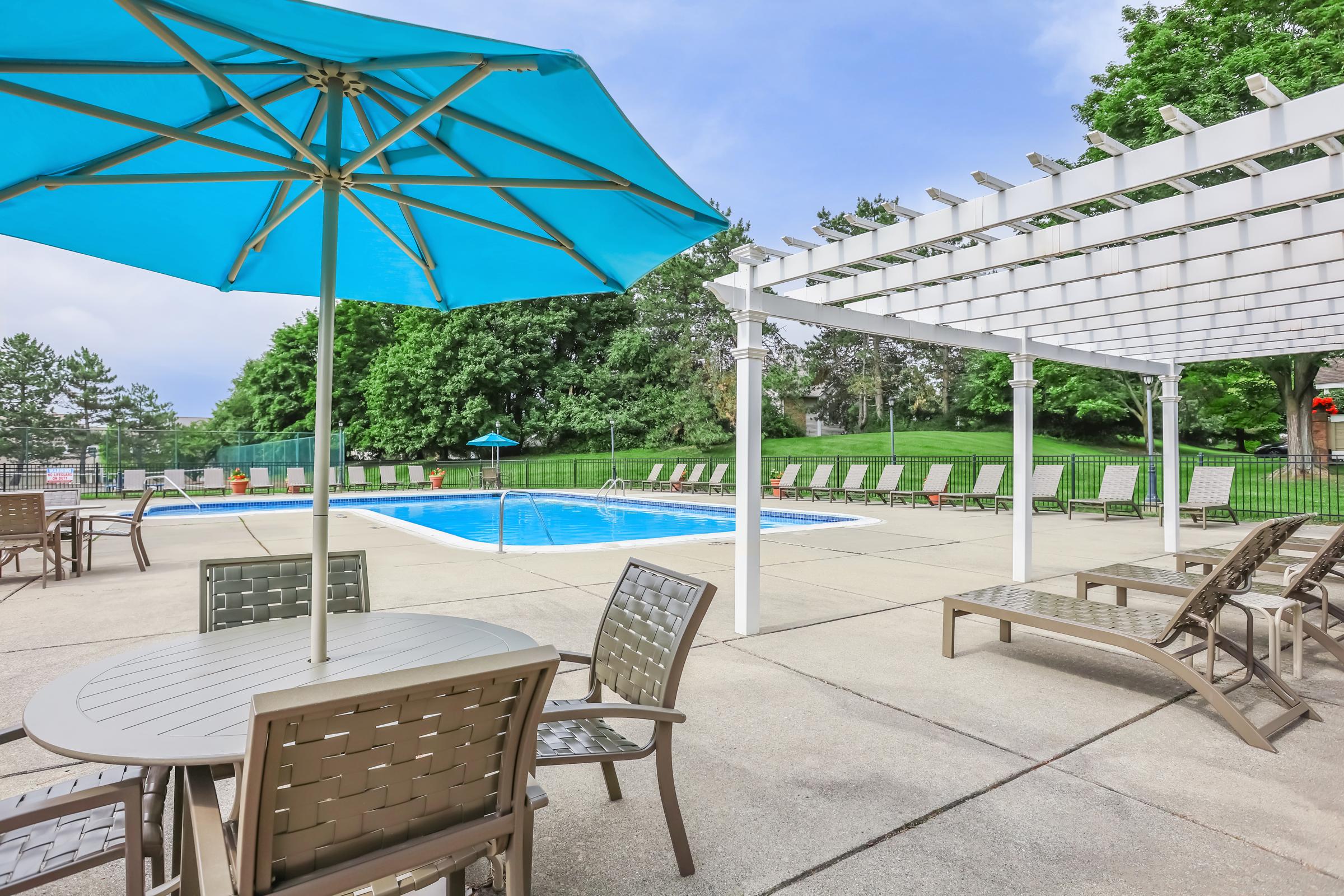 A spacious outdoor pool area featuring a clear blue pool surrounded by lounge chairs and tables. There is a large umbrella providing shade and a pergola nearby, set against a backdrop of lush greenery and a cloudy sky.
