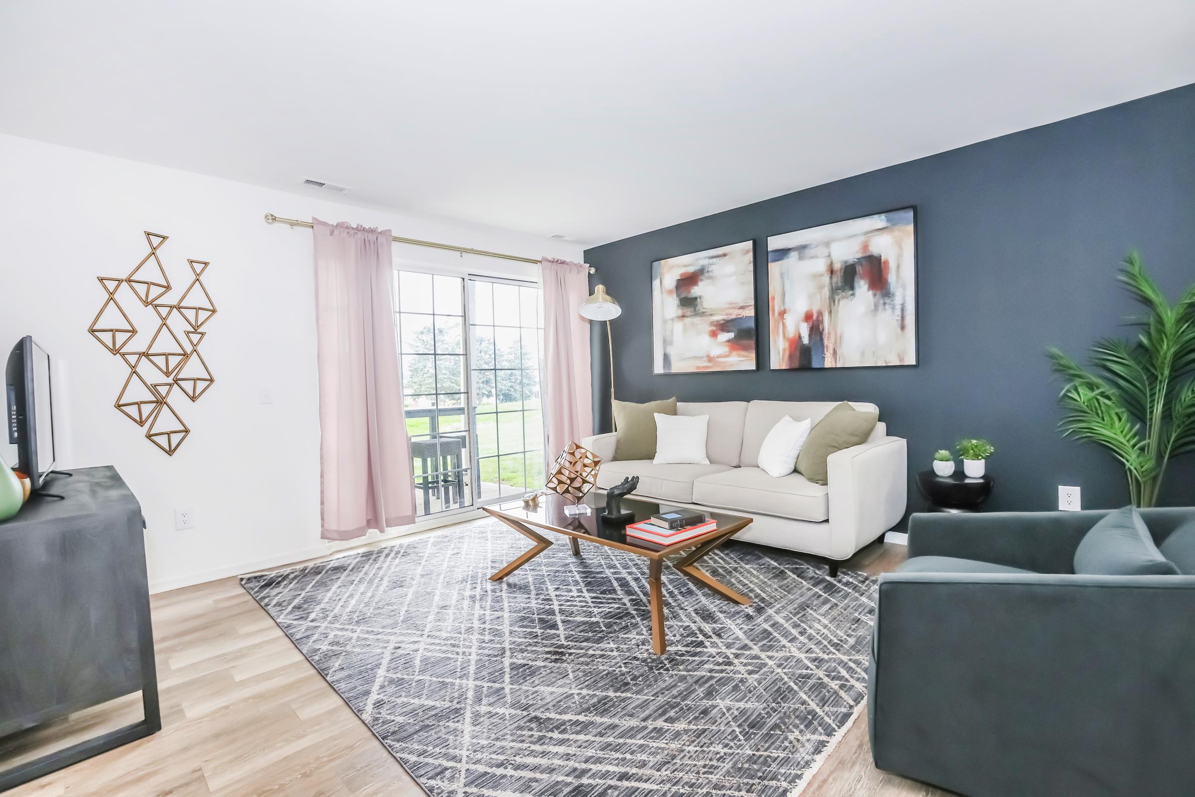 A modern living room featuring a light sofa with green cushions, a stylish coffee table, and an accent chair. Decorative wall art hangs on a dark blue wall, and large windows with sheer curtains let in natural light. A geometric wall decoration and a potted plant add a touch of elegance to the space.