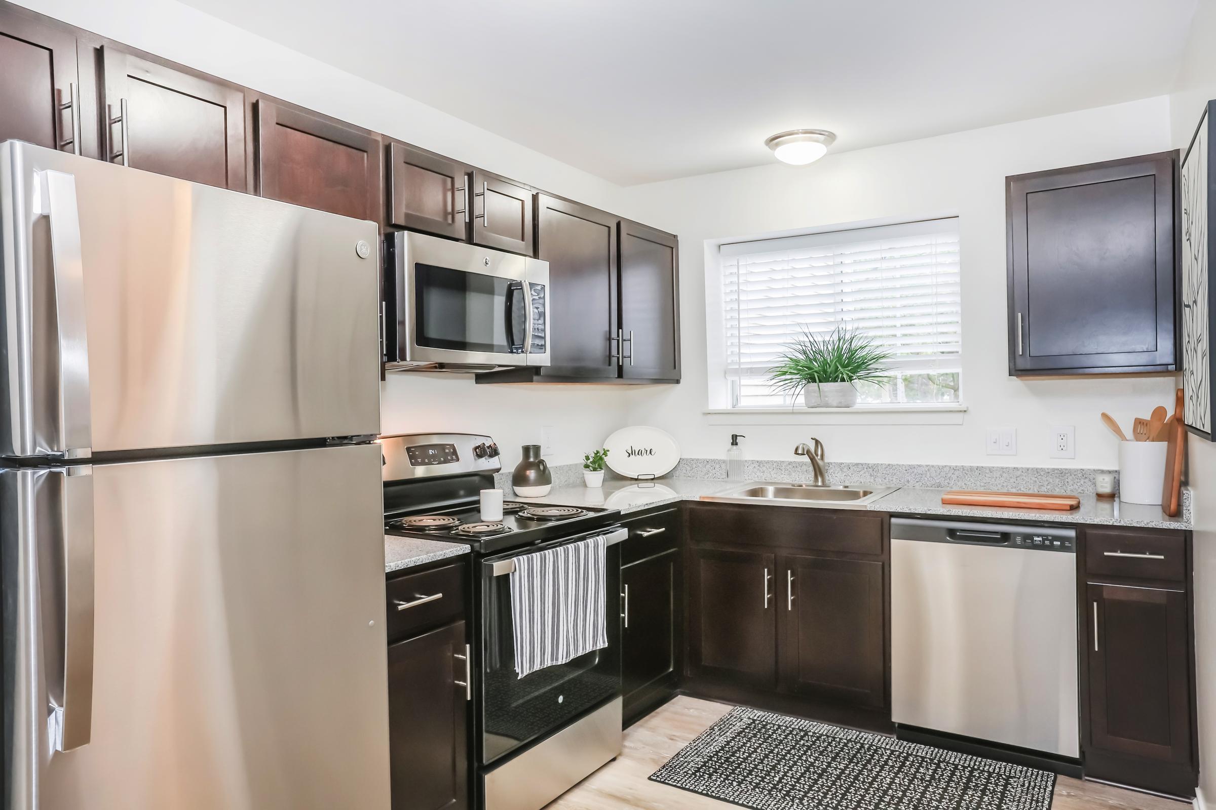 Modern kitchen featuring dark wood cabinets, stainless steel appliances including a refrigerator, microwave, and dishwasher. There's a grey countertop and a window with blinds, along with a small plant on the sill. A black and white striped towel hangs from the oven handle. The overall design is sleek and contemporary.
