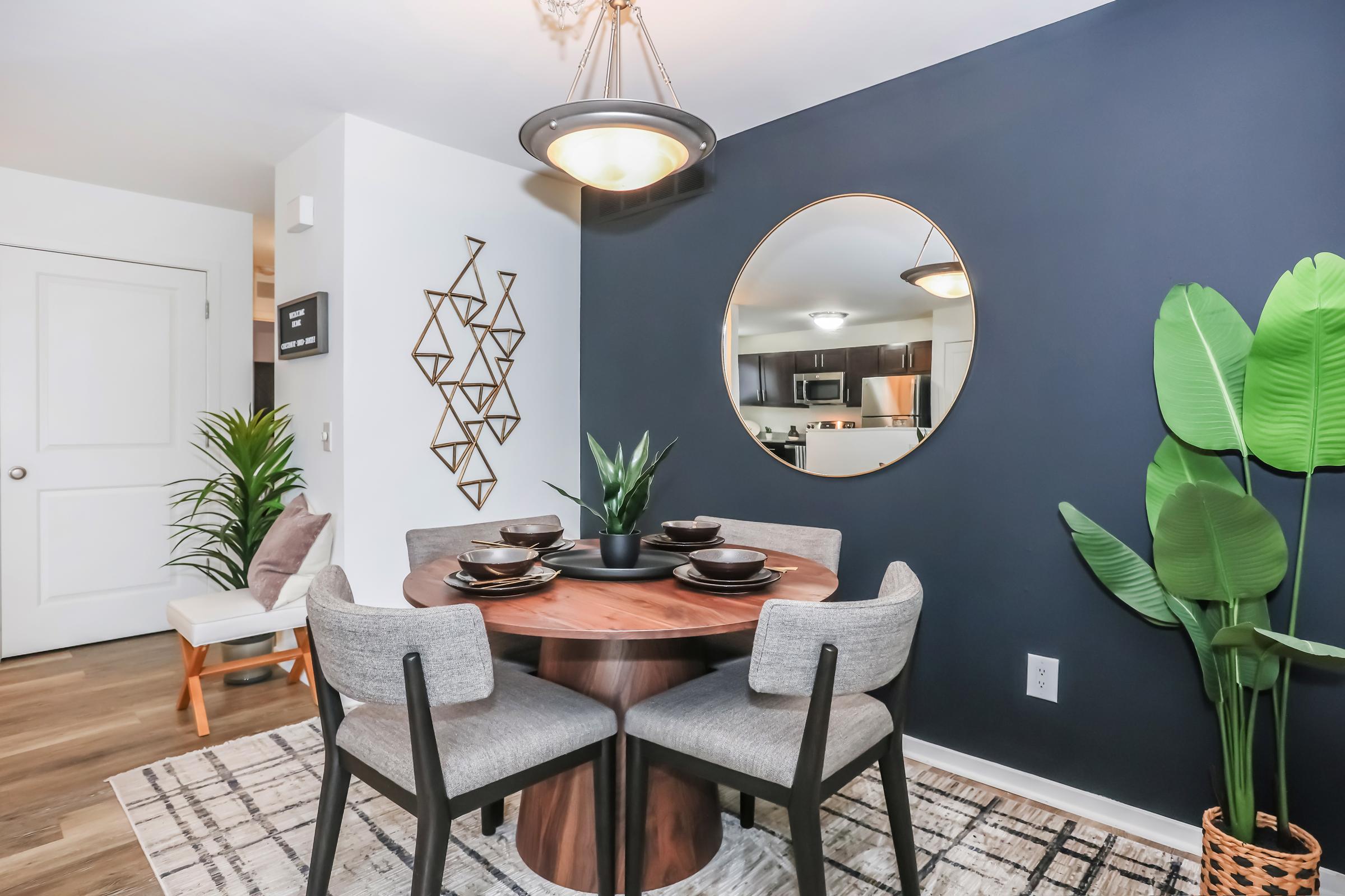 A modern dining area featuring a round wooden table set with black plates and a black centerpiece plant. Gray upholstered chairs surround the table, and a large round mirror hangs on the dark accent wall. Decorative wall art and a potted plant add a touch of greenery, while the space has a contemporary feel.