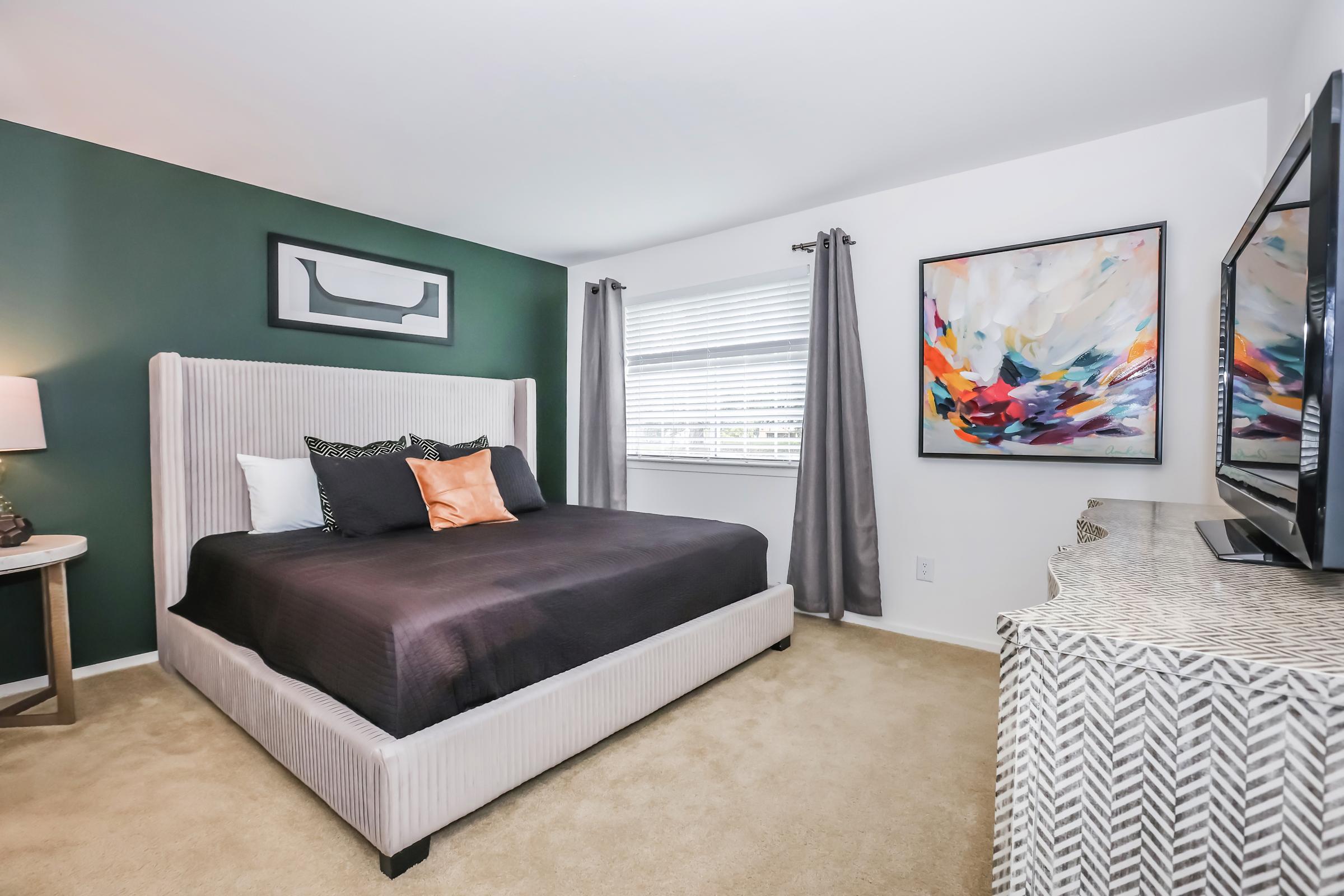 A modern bedroom featuring a beige and black upholstered bed with decorative pillows. The walls are painted dark green and white. A large abstract painting in vibrant colors hangs on one wall, and a sleek dresser sits beside a television. Natural light streams in through the window covered by gray curtains.