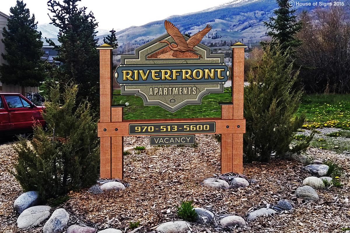 Sign for Riverfront Apartments featuring a stylized eagle. The sign includes the phone number "970-513-5600" and indicates availability with "VACANCY." Surrounding the sign are landscaped rocks and plants, set against a backdrop of mountains and trees.