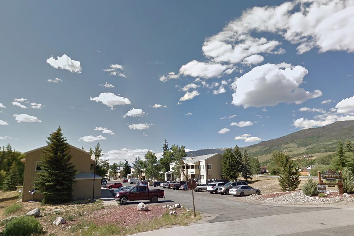 Street view of a small town with residential buildings and parked cars. Lush green trees line the road, and the background features rolling hills under a partly cloudy sky with scattered clouds. The scene conveys a quiet, suburban atmosphere.