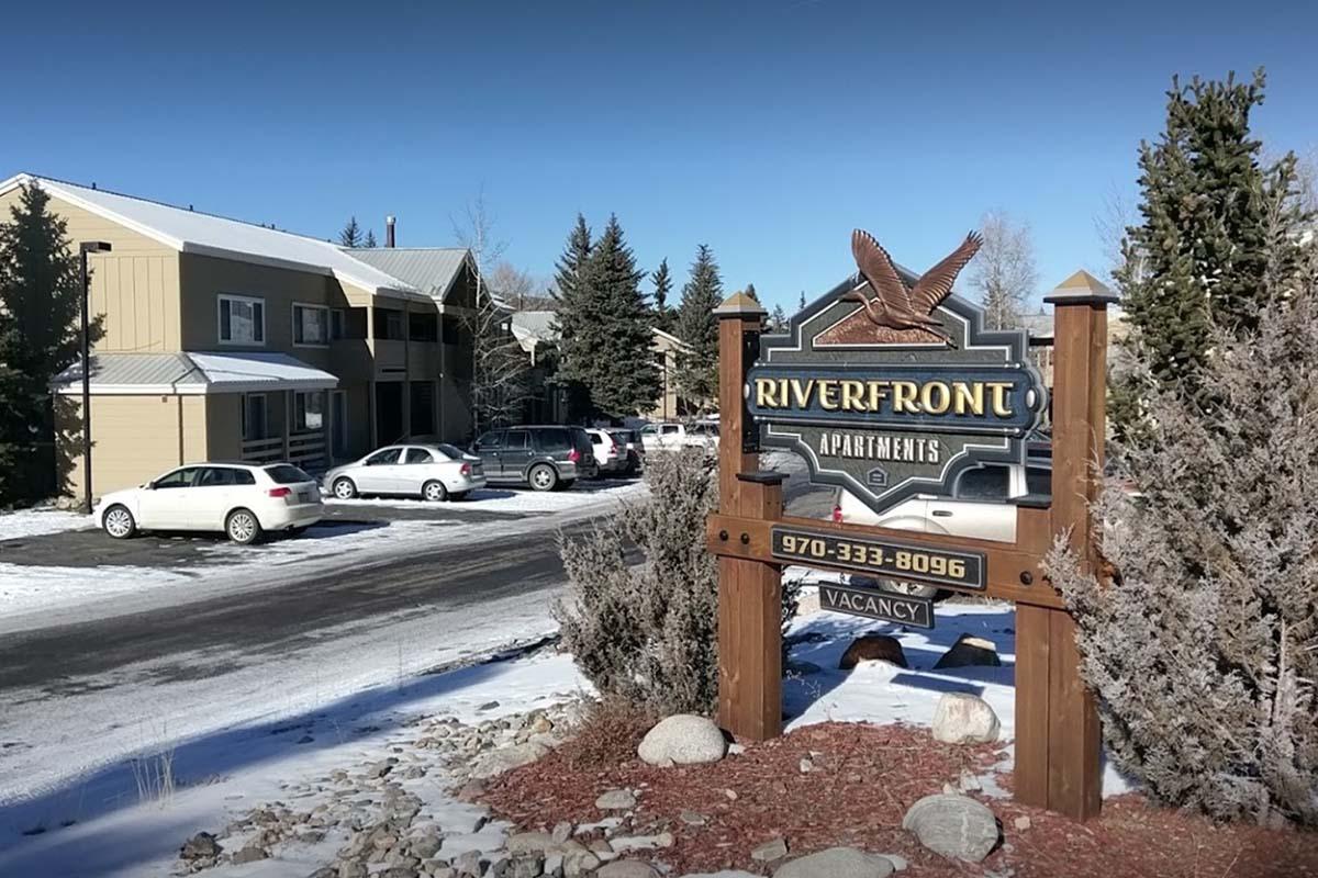 Sign for Riverfront Apartments with a stone base and wooden details, featuring an eagle emblem. Surrounding the sign are snowy landscapes, tall trees, and parked cars. In the background, there's a building with several apartments visible. The sky is clear and blue.