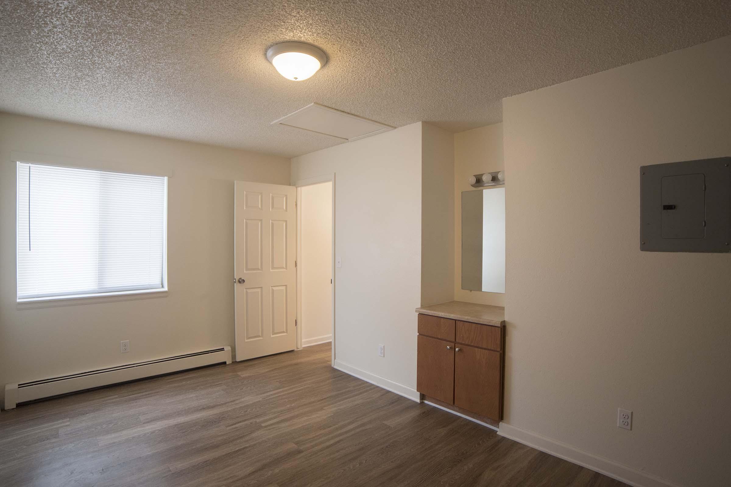 A spacious, well-lit living area with light-colored walls and a wooden floor. It features a window with blinds, a closet door, and a small mirror above a wooden cabinet. The ceiling has a simple light fixture, and there is an electrical panel on the wall. Overall, the room has a clean and modern appearance.