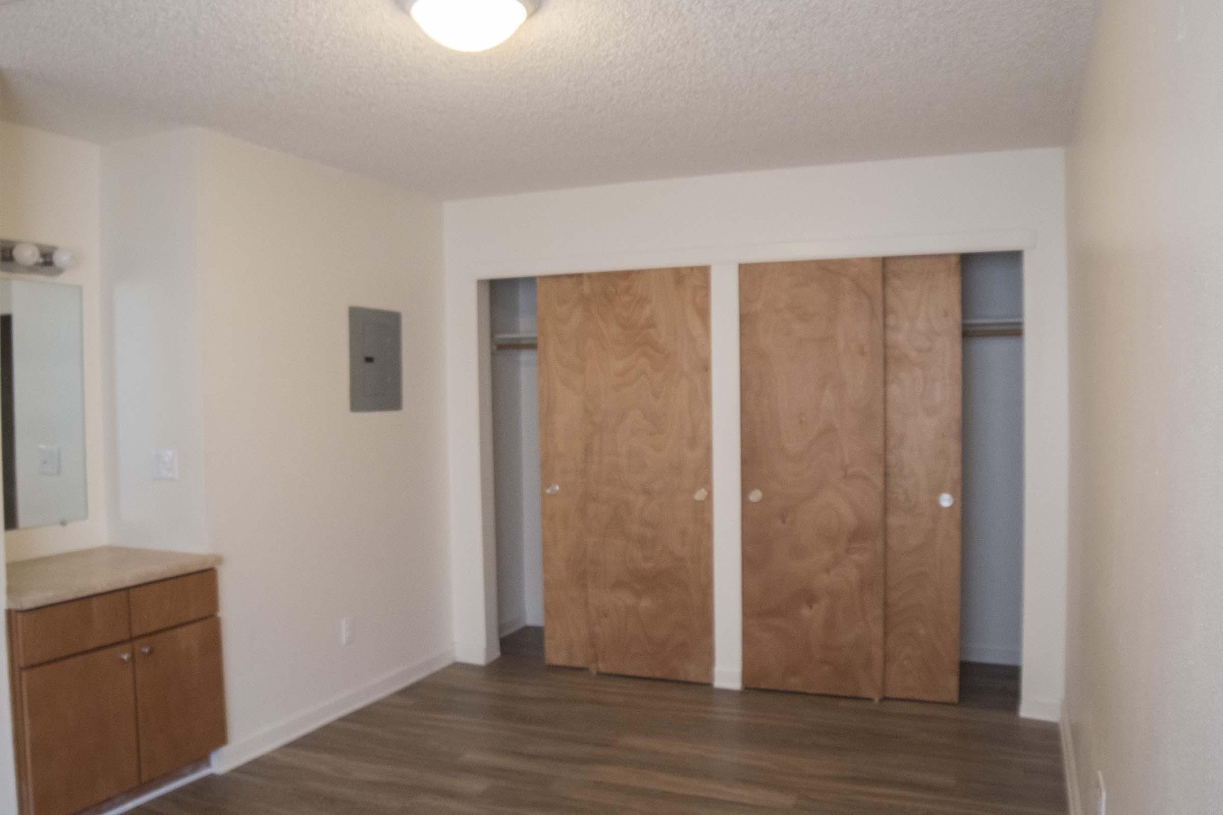 A simple, empty room featuring a light fixture, a small vanity with a mirror, and two wooden closet doors. The walls are painted a light color, and the floor is covered with laminate flooring. The room appears well-lit and spacious.