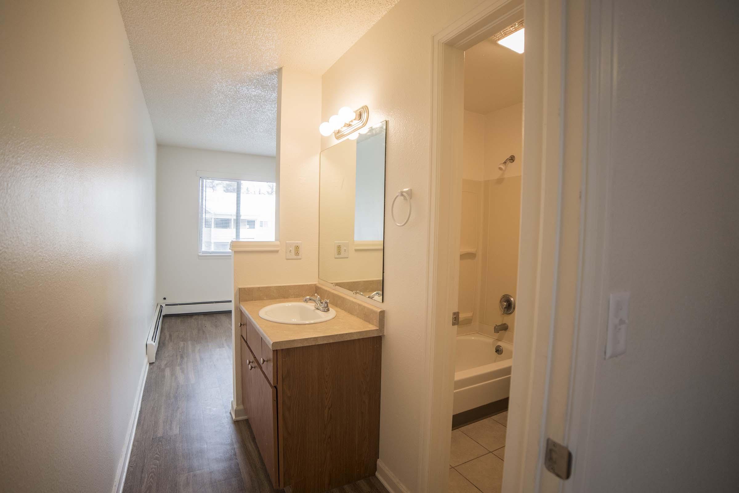 A small bathroom with a sink and mirror on a counter, and a bathtub/shower combination visible. The adjacent room is empty with natural light coming from a window, and there is a wooden floor throughout. The walls are painted light colors, creating a clean and spacious appearance.