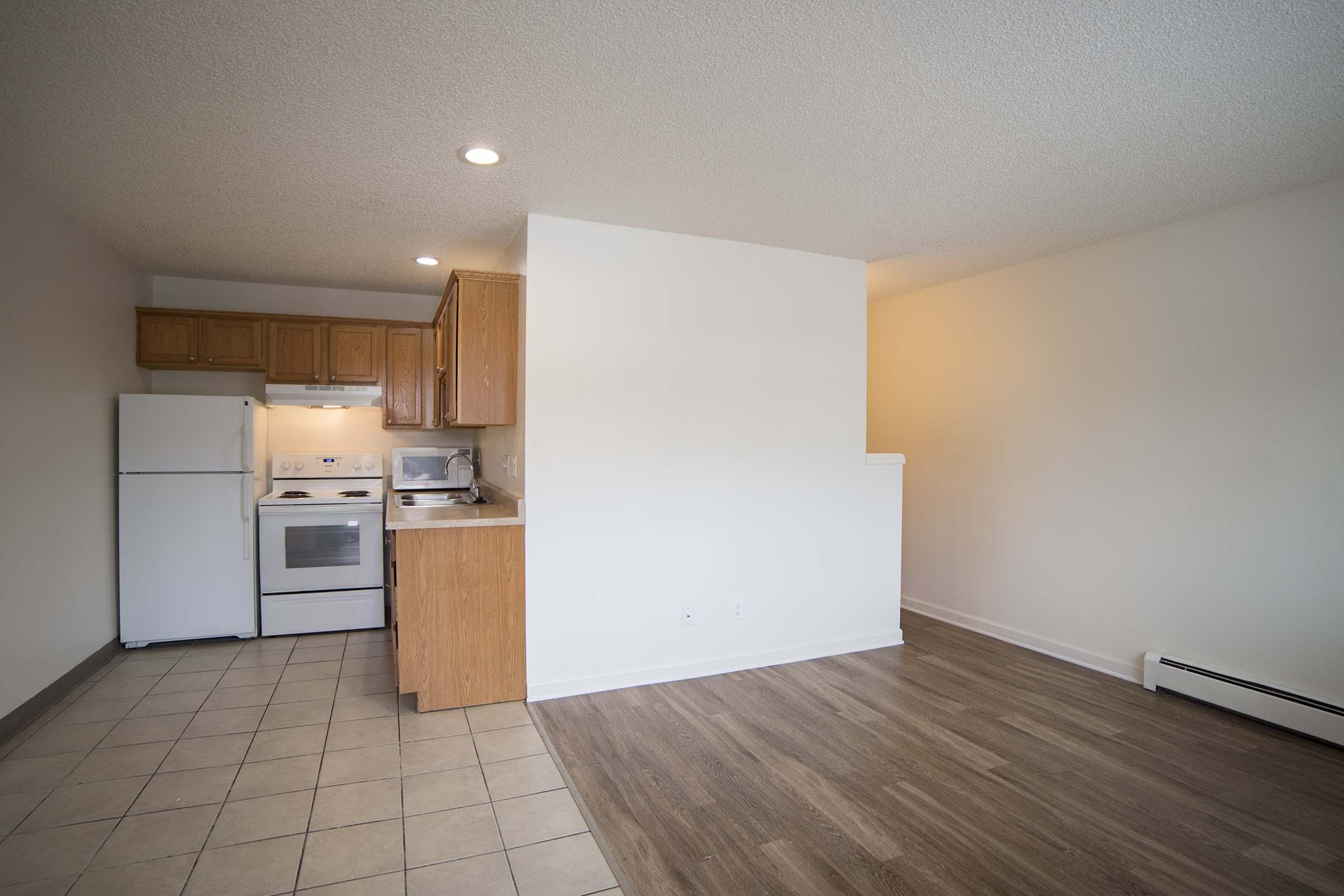 An open-concept living area featuring a kitchen with wooden cabinets, white appliances, and a tiled floor. The adjacent space has light-colored walls and a wooden floor, providing a bright and airy ambiance. The layout allows for easy movement between the kitchen and living area.