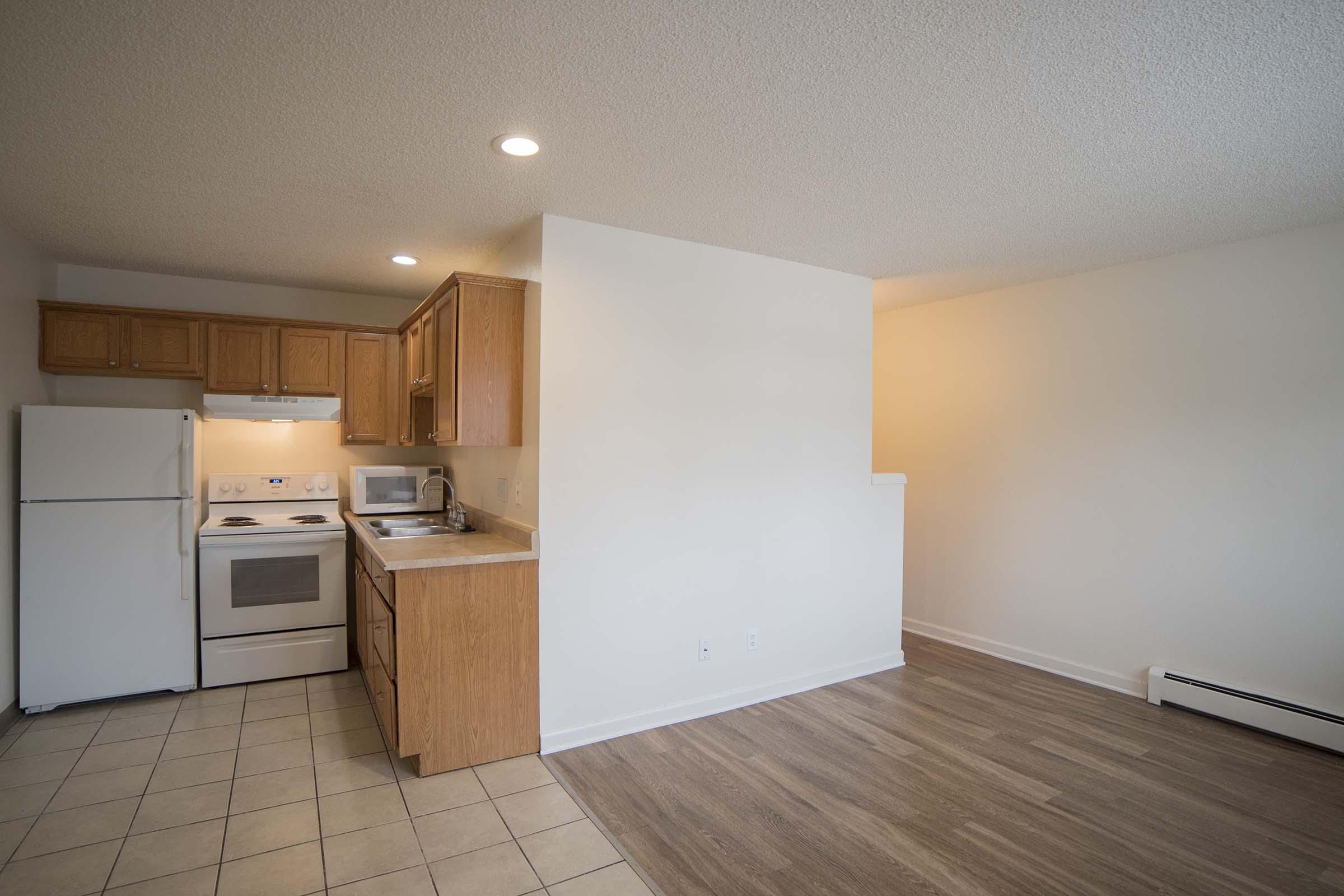 A modern apartment kitchen featuring wooden cabinets, a white refrigerator, oven, and microwave. The open layout connects to a light-filled living area with neutral walls and laminate flooring, creating a spacious feel.