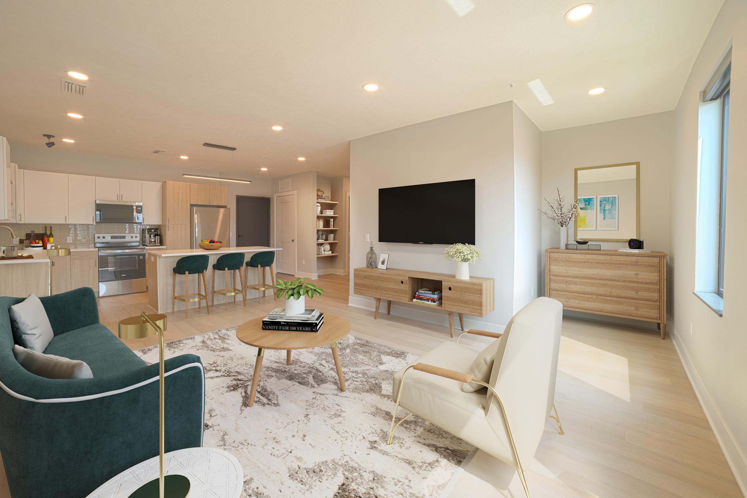 A modern living room featuring a comfortable seating area with a green sofa and a stylish white chair. There's a round coffee table in the center, and a large area rug beneath. The space includes a kitchen in the background with barstool seating, and a TV mounted on the wall, complemented by a decorative cabinet.