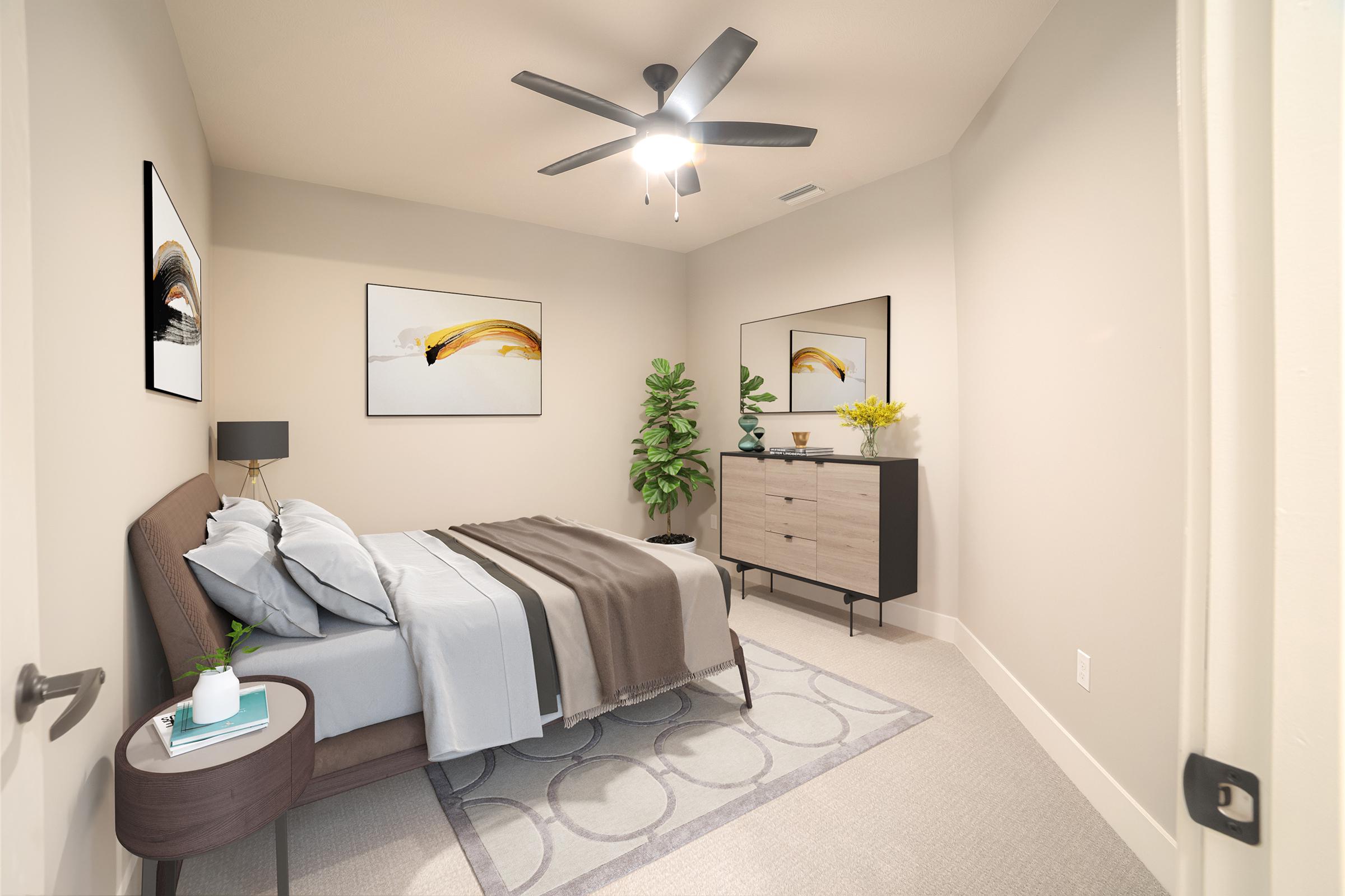 A cozy bedroom featuring a bed with light blue bedding and decorative pillows, a bedside table with a small plant, and a modern dresser. Two abstract paintings adorn the walls, and a potted plant adds a touch of greenery. A ceiling fan provides airflow, enhancing the inviting atmosphere.