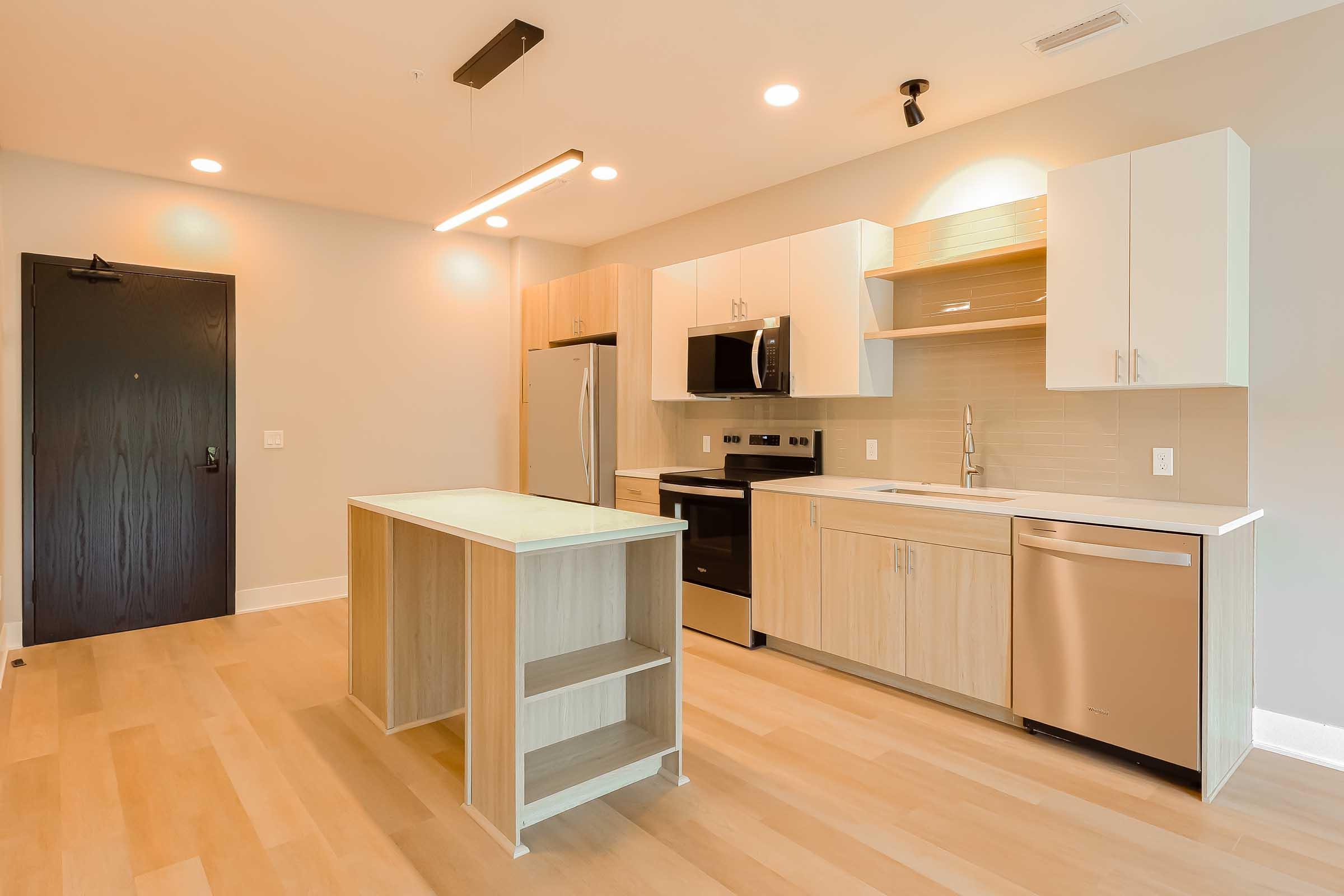 Modern kitchen featuring light wood cabinetry, stainless steel appliances including a refrigerator, oven, and dishwasher. A central island with an open shelf stands in the middle, with white countertops and pendant lighting above. The flooring is light-colored wood, and the walls are neutral-toned.