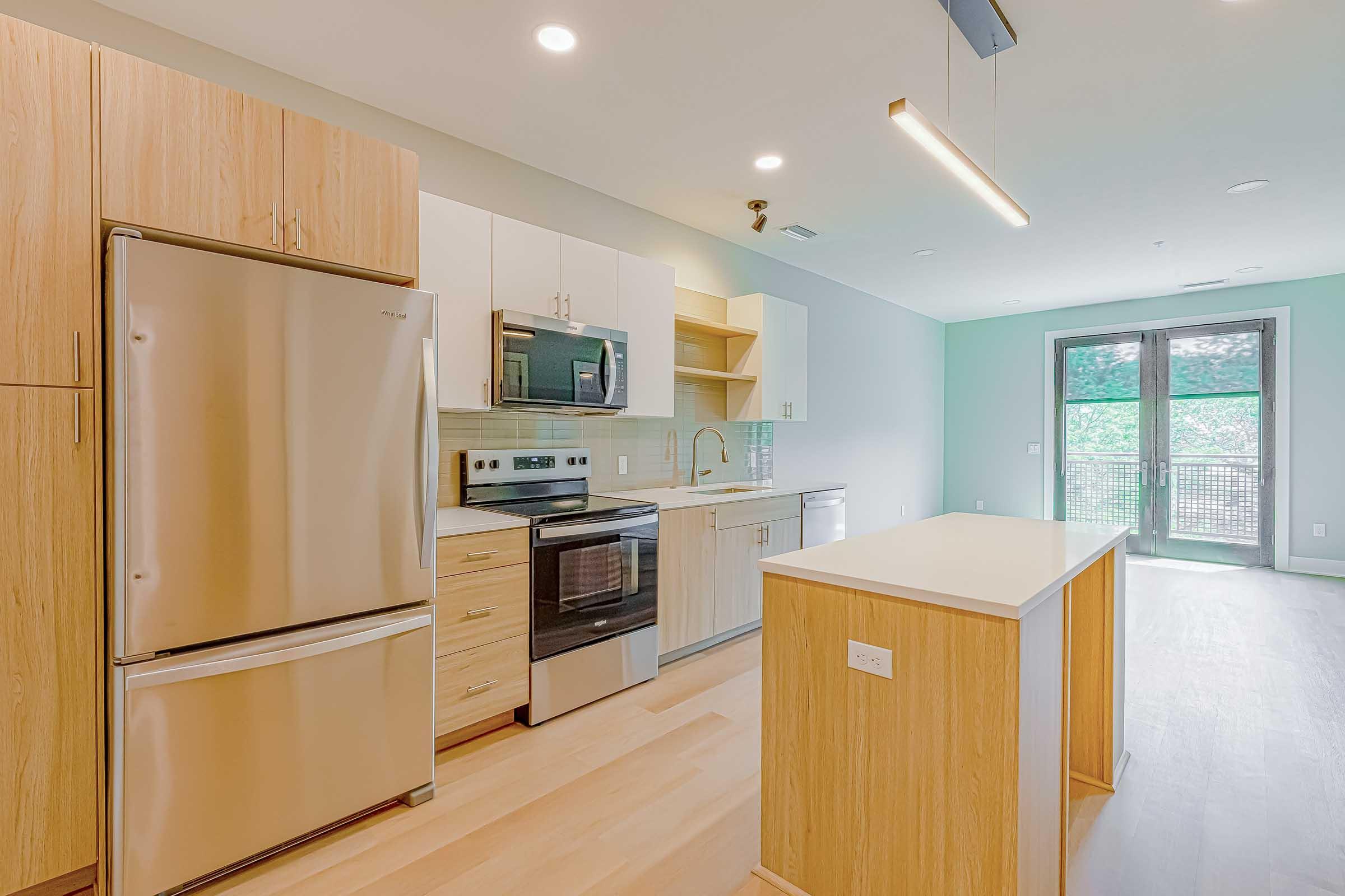 Modern kitchen featuring stainless steel appliances, including a refrigerator and oven, light wood cabinetry, and a spacious island. The walls are light-colored, with one wall painted in a soft mint green. Natural light comes in through a sliding glass door leading to a patio or balcony.