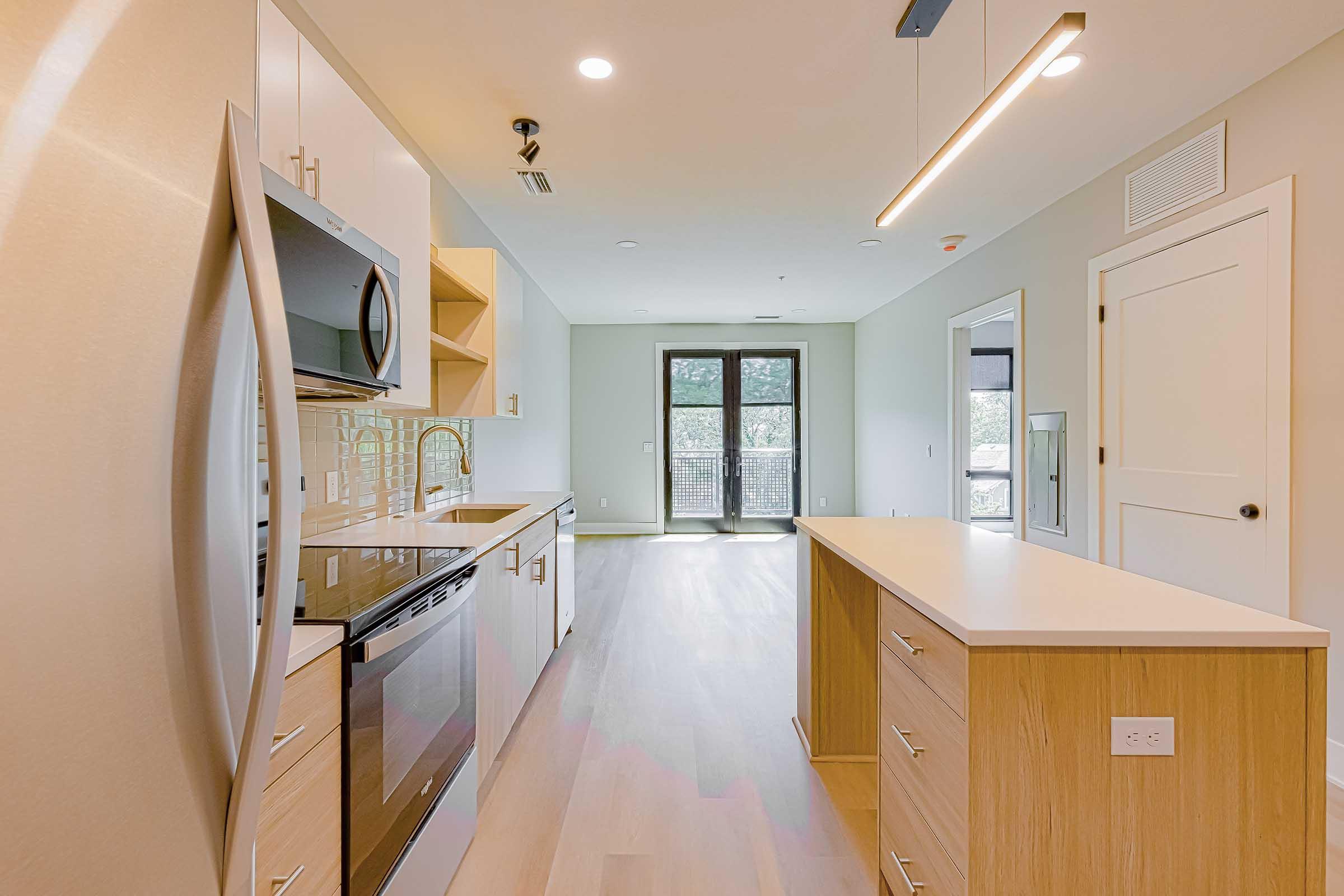 Modern kitchen featuring stainless steel appliances, including a refrigerator and oven. There's a large island with a countertop, light wood cabinets, and a sliding glass door leading to an outdoor area. The walls are painted a soft neutral color, creating a bright, airy atmosphere.
