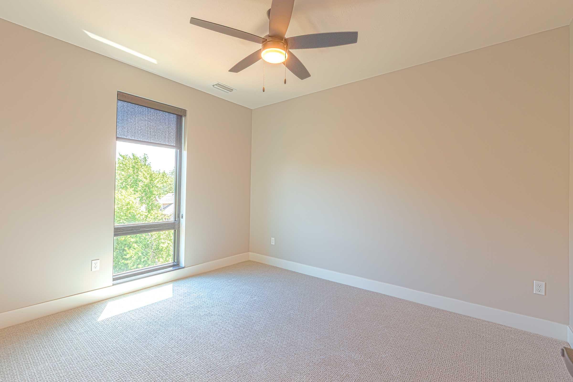 A spacious, empty room with light beige walls and a light-colored carpet. A ceiling fan is mounted on the ceiling, and natural light streams in through a large window covered with a roller shade. The overall atmosphere is bright and airy.