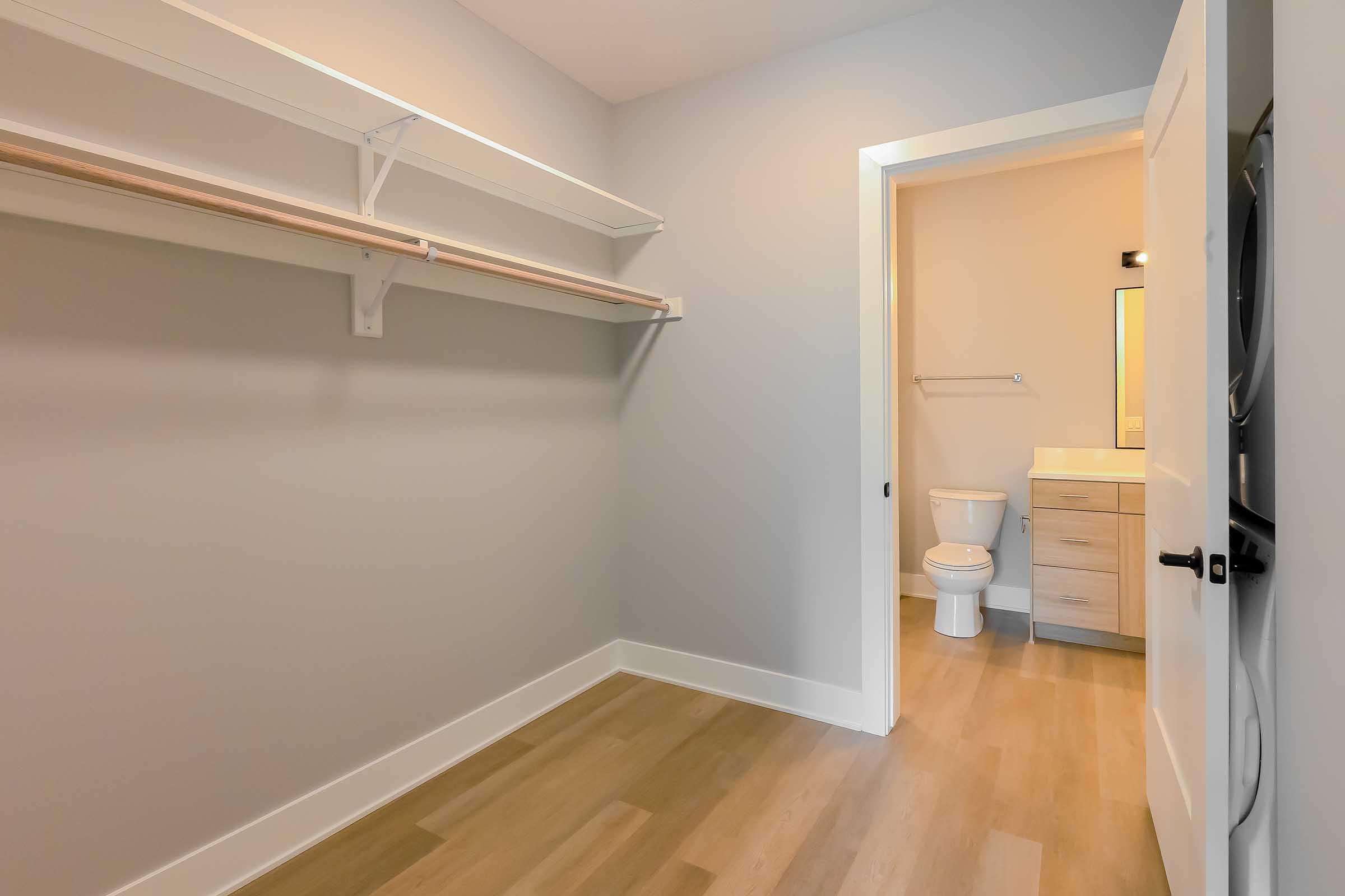 A minimalistic room featuring light gray walls, a wooden floor, and a small bathroom area visible through an open door. There are shelves on one wall, and a bathroom with a toilet and a vanity is visible in the background. The space appears bright and well-lit, with natural light coming in.