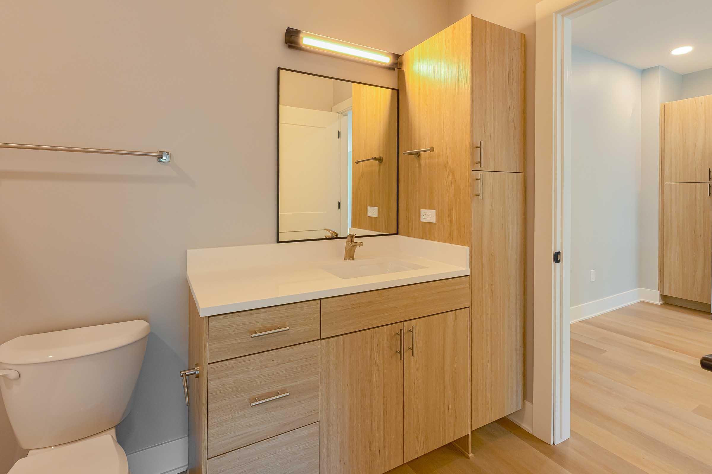 Modern bathroom featuring a wooden vanity with a sink and faucet, a large mirror, wall-mounted light fixture, and a toilet. The floors are light-colored wood, and the walls are painted in a soft neutral tone. There are minimalistic cabinets for storage, creating a clean and contemporary aesthetic.