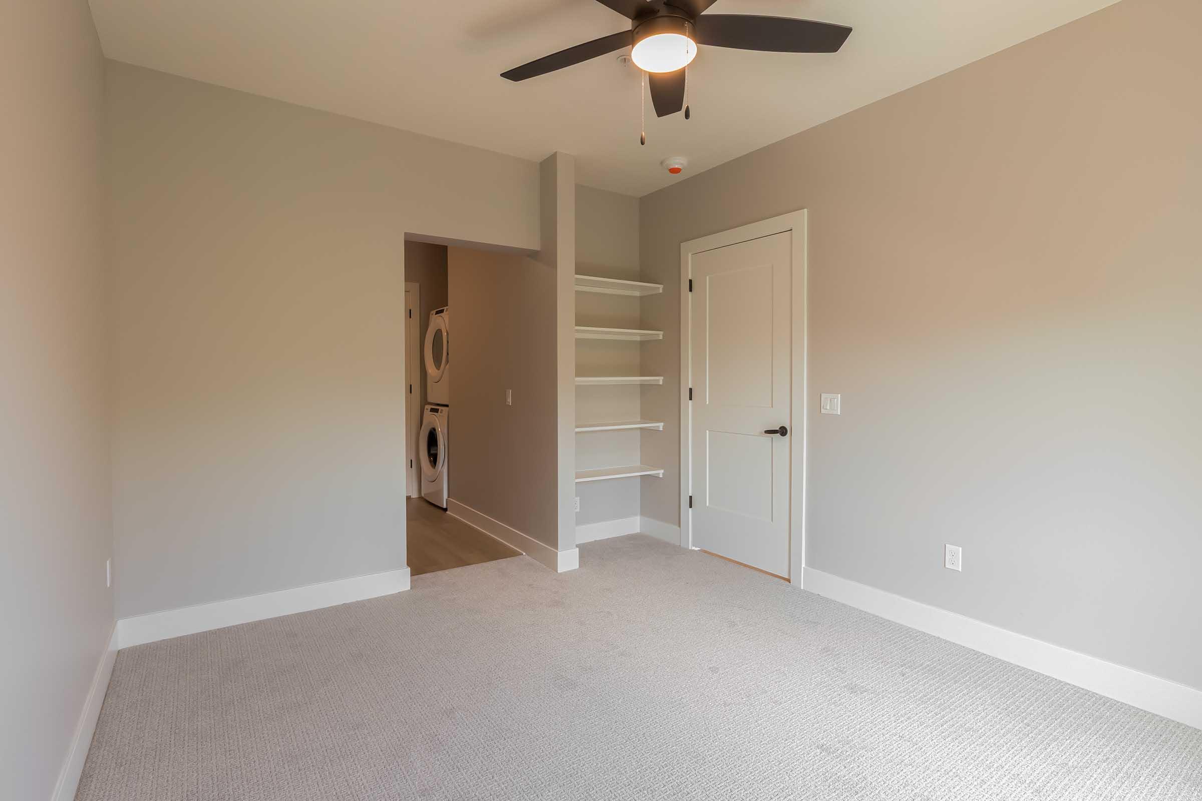 A vacant room featuring light gray walls, carpeted floor, and a ceiling fan. There are open shelves against one wall and a doorway leading to another space, which appears to contain laundry appliances. The room is well-lit with natural light.