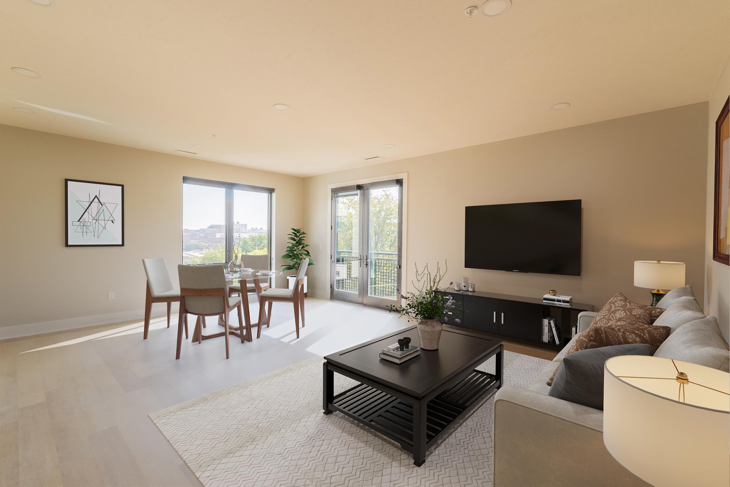 A bright and modern living room with light-colored walls and wooden flooring. It features a comfortable gray sofa, a black coffee table, a flat-screen TV on a dark cabinet, and a small dining area with four chairs. Large windows provide natural light and showcase greenery outside. A plant adds a touch of decor.