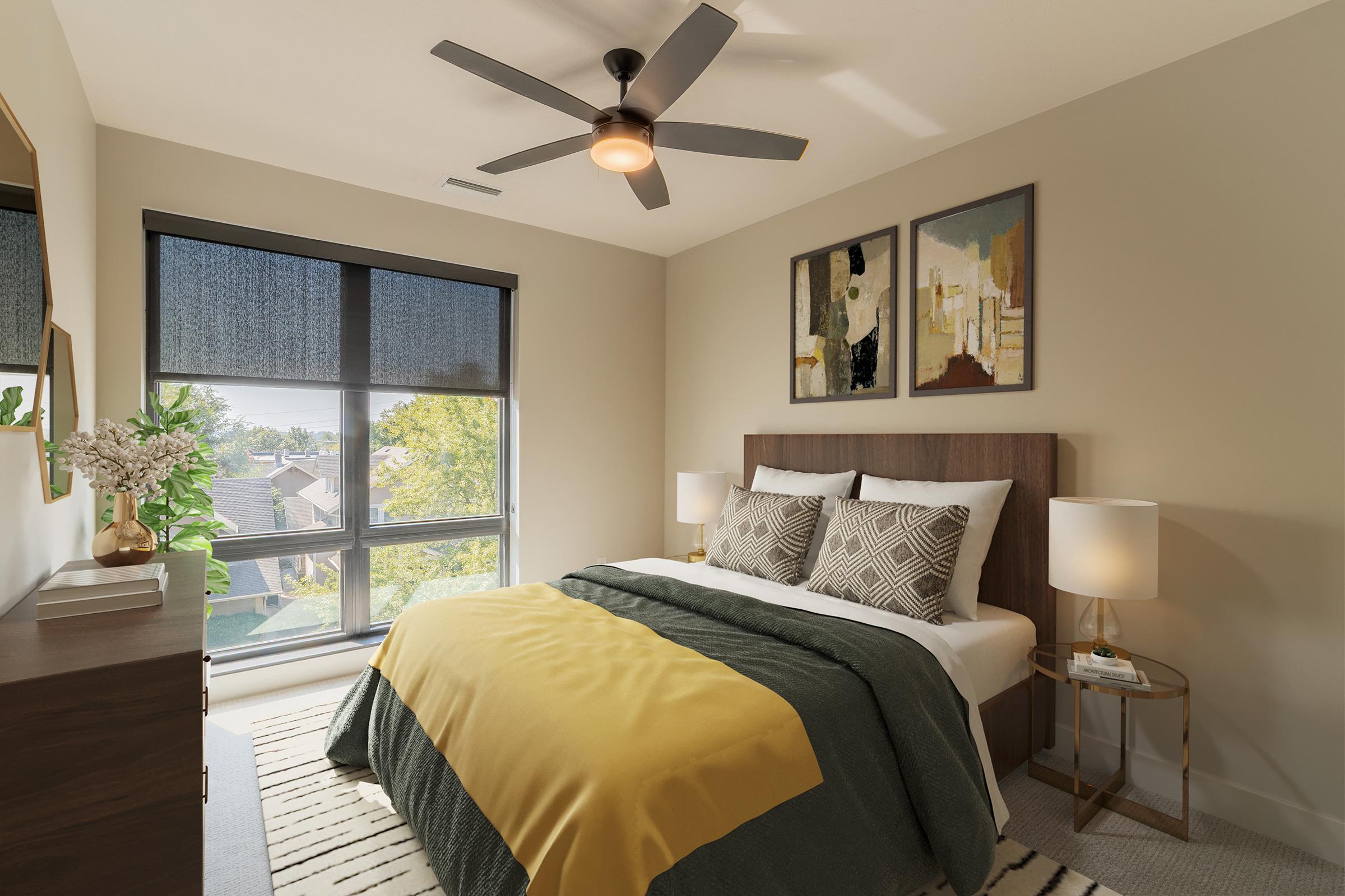 A well-decorated bedroom featuring a queen-sized bed with patterned pillows and a yellow throw blanket. There's a wooden headboard, a stylish nightstand with a lamp, and a dresser. The large window provides natural light, and artwork hangs on the wall. A cozy rug adds warmth to the space.