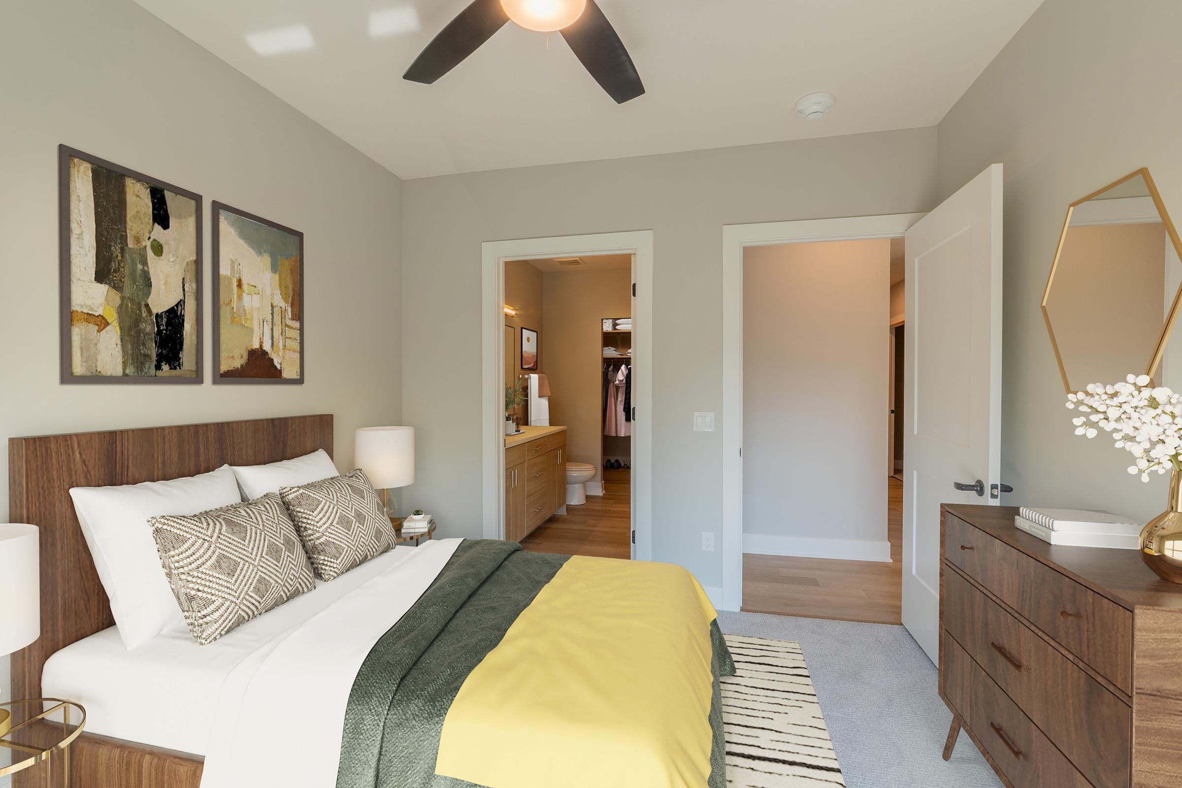A cozy bedroom featuring a modern design with a wooden bed frame dressed in white and green bedding. Two abstract paintings hang above the bed, and a dresser is placed beside it. A door leads to a bathroom area, and natural light fills the room through an open door. A ceiling fan is visible above.