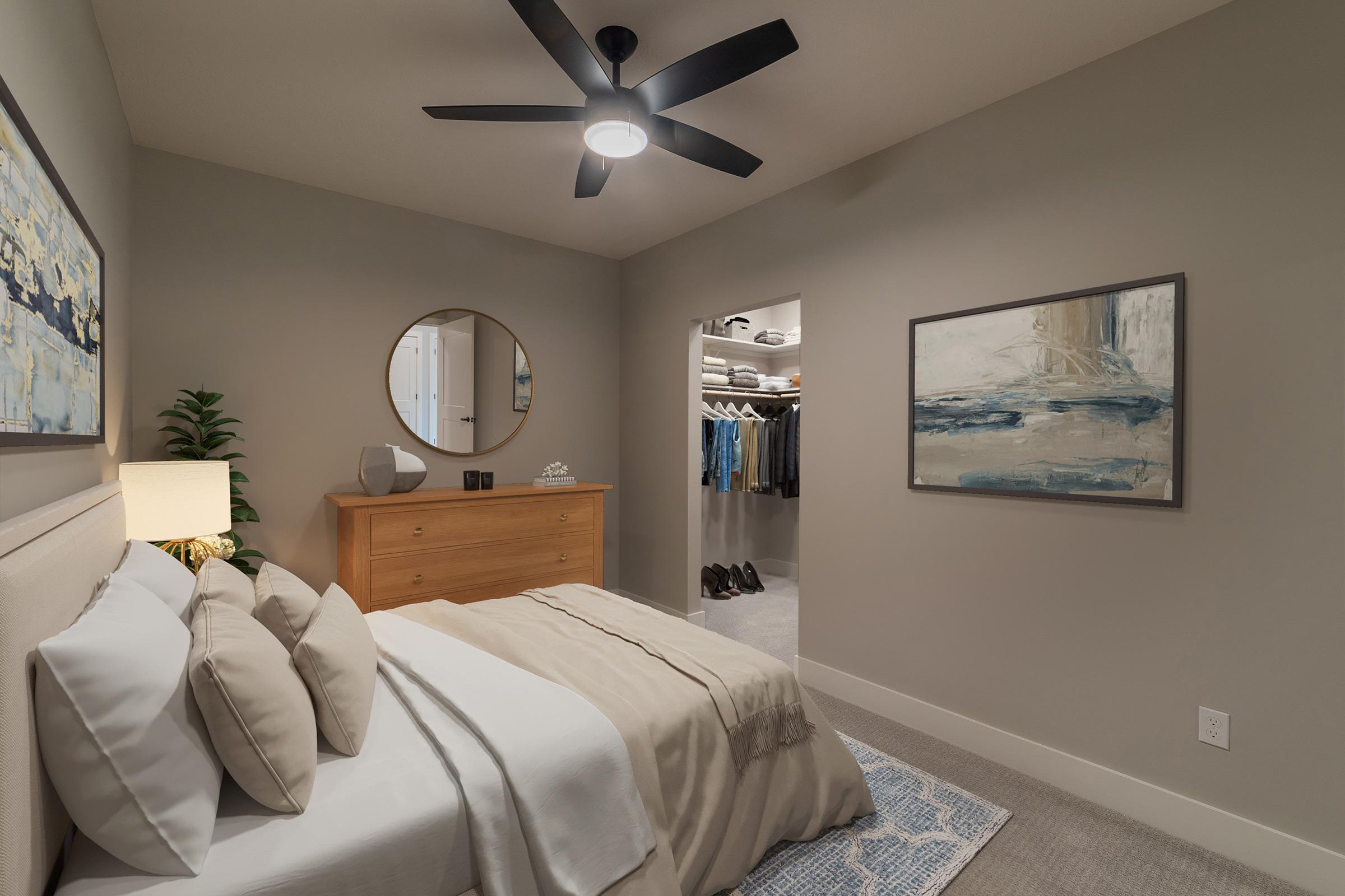 Cozy bedroom featuring a large bed with soft, neutral bedding, a stylish dresser, a round mirror, and a ceiling fan. A small plant adds a touch of greenery, while a closet in the background displays neatly hung clothing. The artwork on the wall complements the overall calming color scheme.