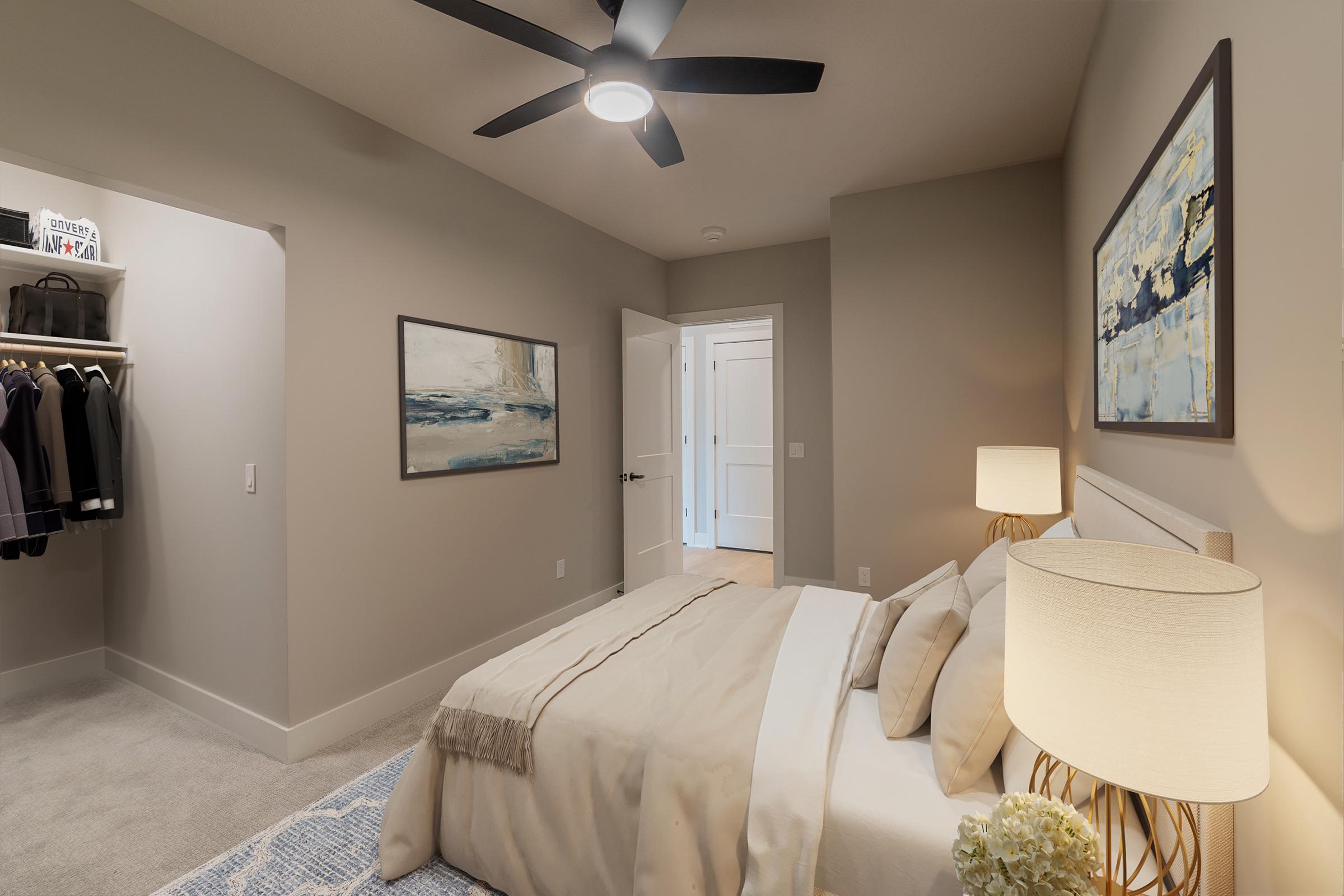 A cozy bedroom featuring a neatly made bed with a light-colored duvet, a bedside lamp, and a modern ceiling fan. The walls are painted in soft tones, and artwork hangs above the bed. A door leads to a closet with neatly arranged clothing, while another door opens to a light-filled hallway.