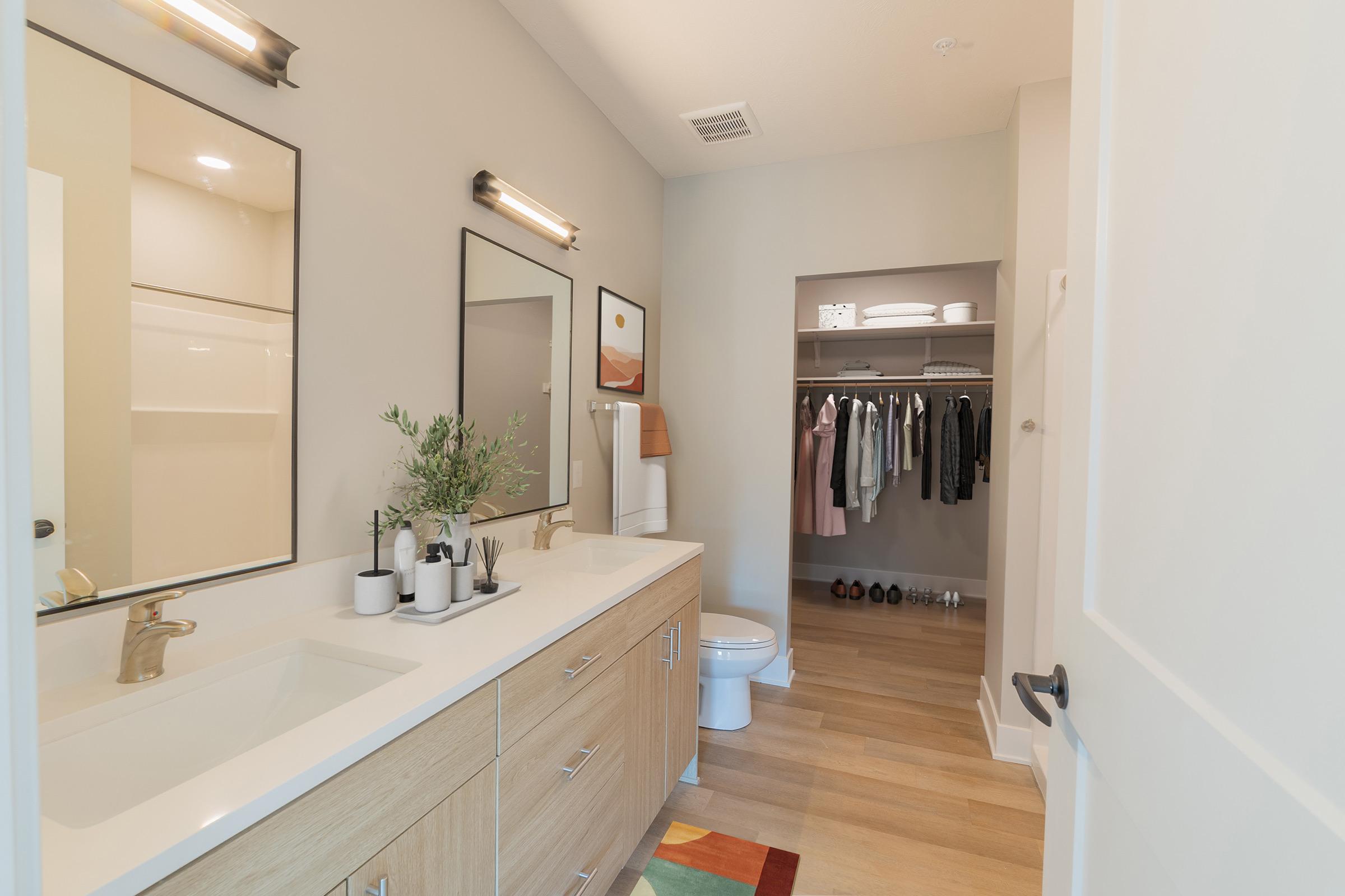 A modern bathroom featuring a double sink with a light wood vanity, two mirrors, and stylish lighting. In the background, there's a neatly organized walk-in closet with hanging clothes and a few pairs of shoes visible. The floor is light wood, and a colorful rug adds a decorative touch.