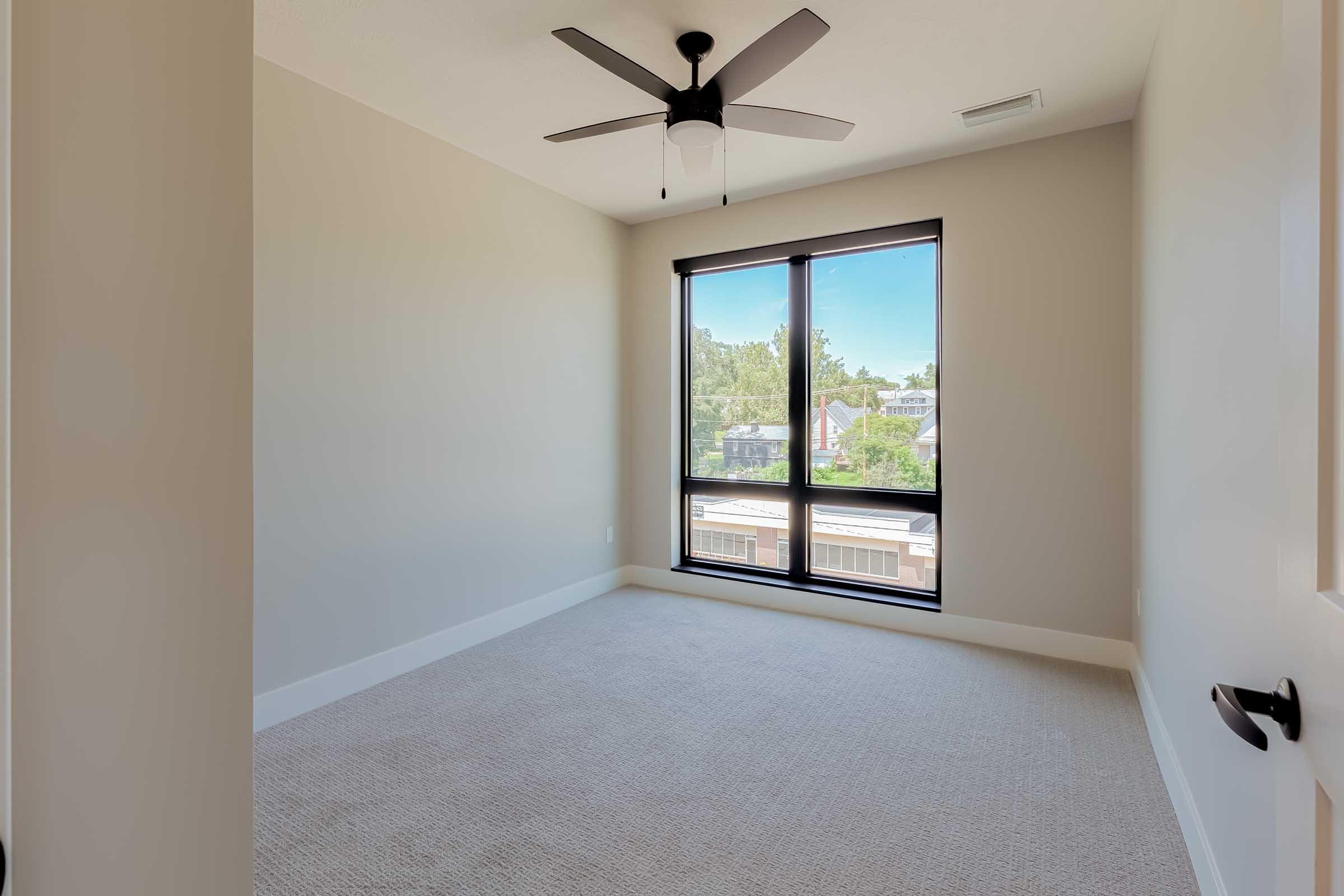 A bright, empty bedroom featuring light beige walls and a large window with black frames. The floor is covered in light carpet, and a modern ceiling fan hangs from the ceiling. The room has a minimalist design with no furniture, creating an open and airy feel.