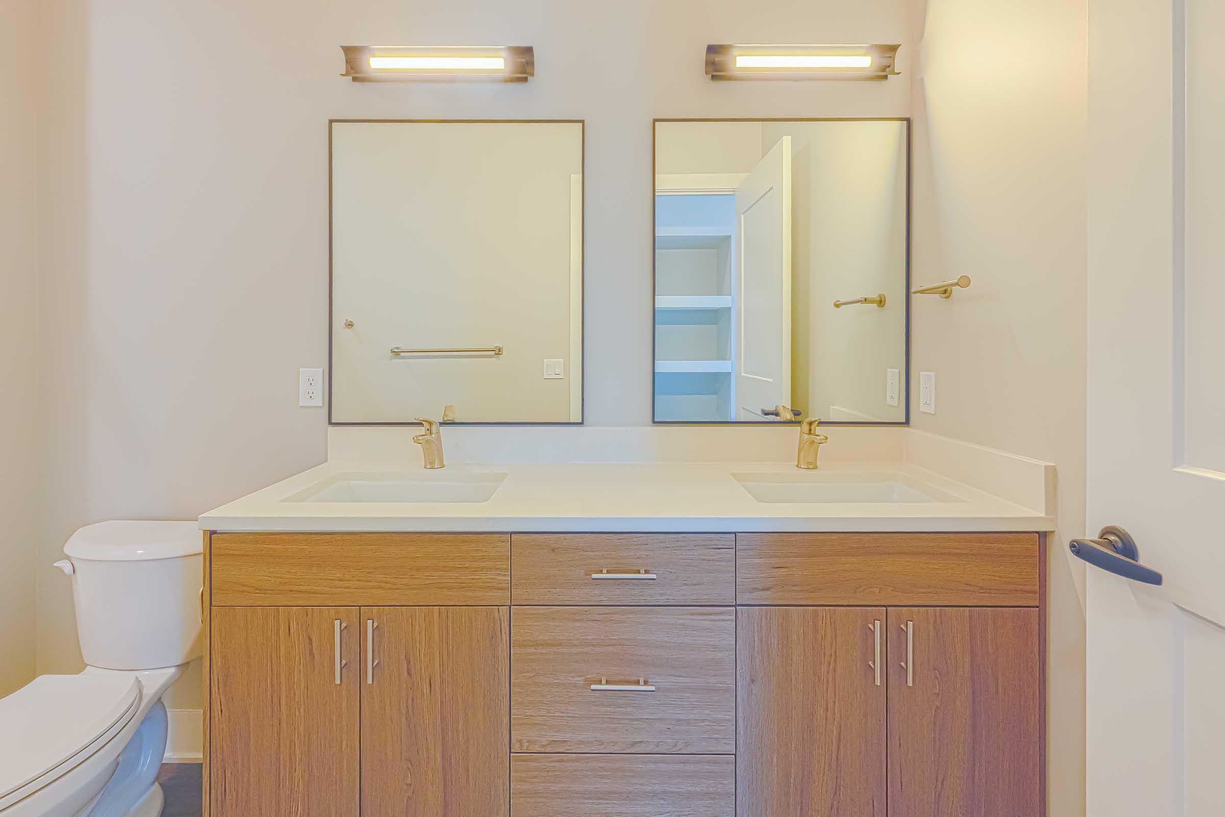 A modern bathroom featuring a double vanity with two sinks, bronze faucets, and two mirrors above. Light-colored walls and a stylish door are visible, along with a white toilet and a storage cabinet in the background. The overall design is clean and contemporary.