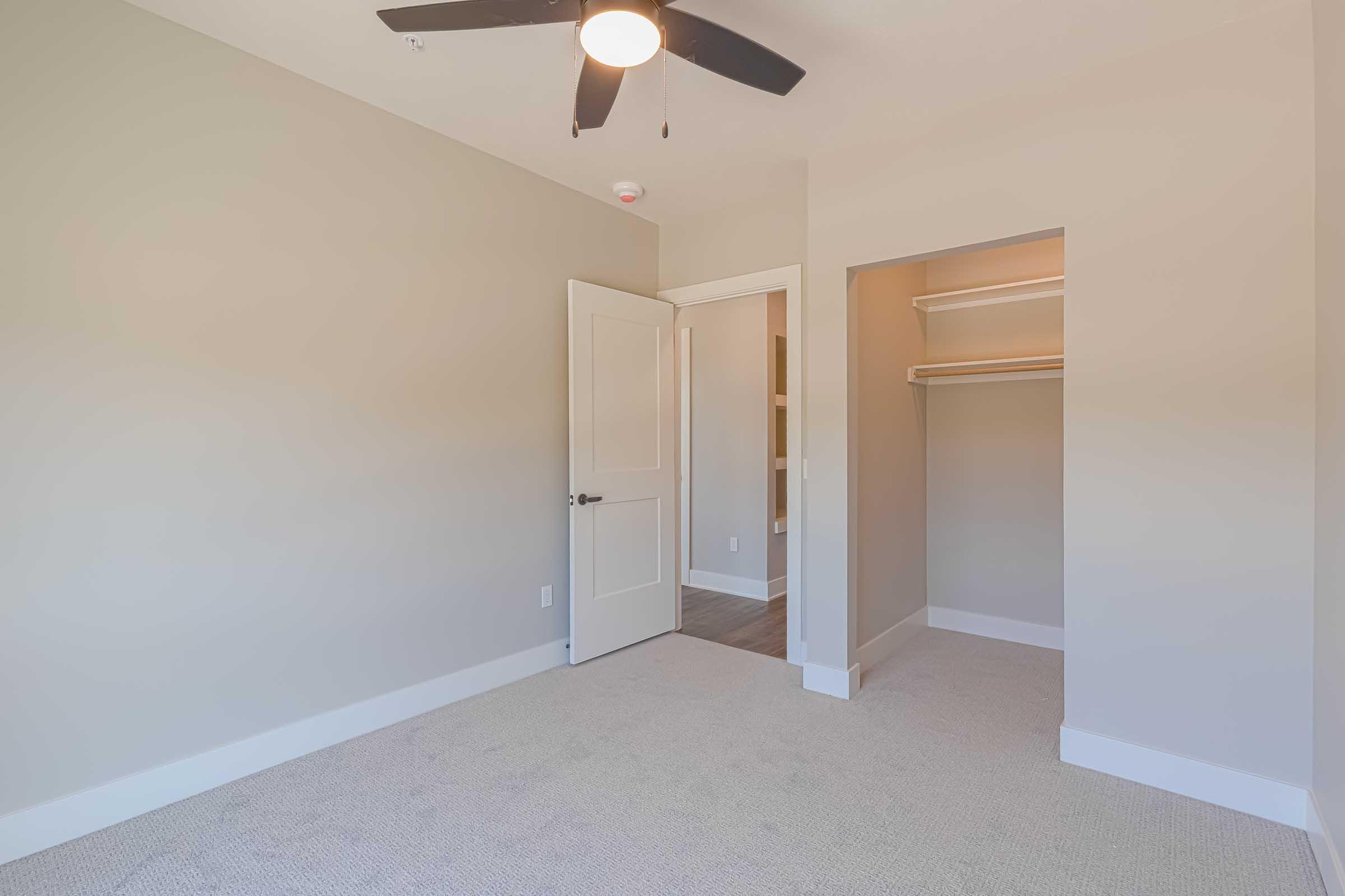 A well-lit, empty room with a ceiling fan, beige walls, and light carpet. There's a closed door on the left leading to another space, and an open closet on the right against the wall. The room has a modern feel with simple architectural details, perfect for personal design.